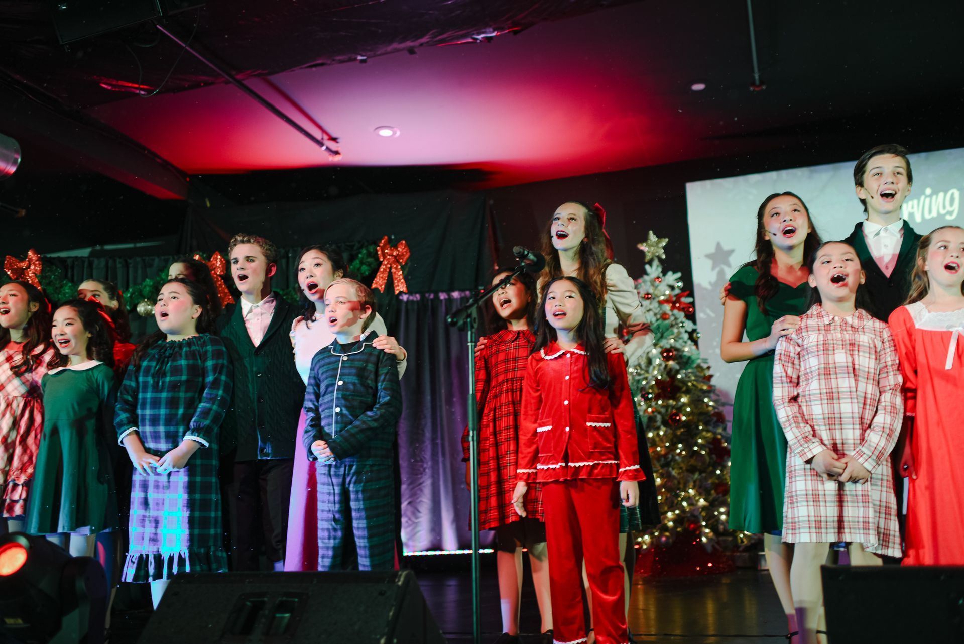 Children singing on a stage in colorful outfits with red and white lights behind them