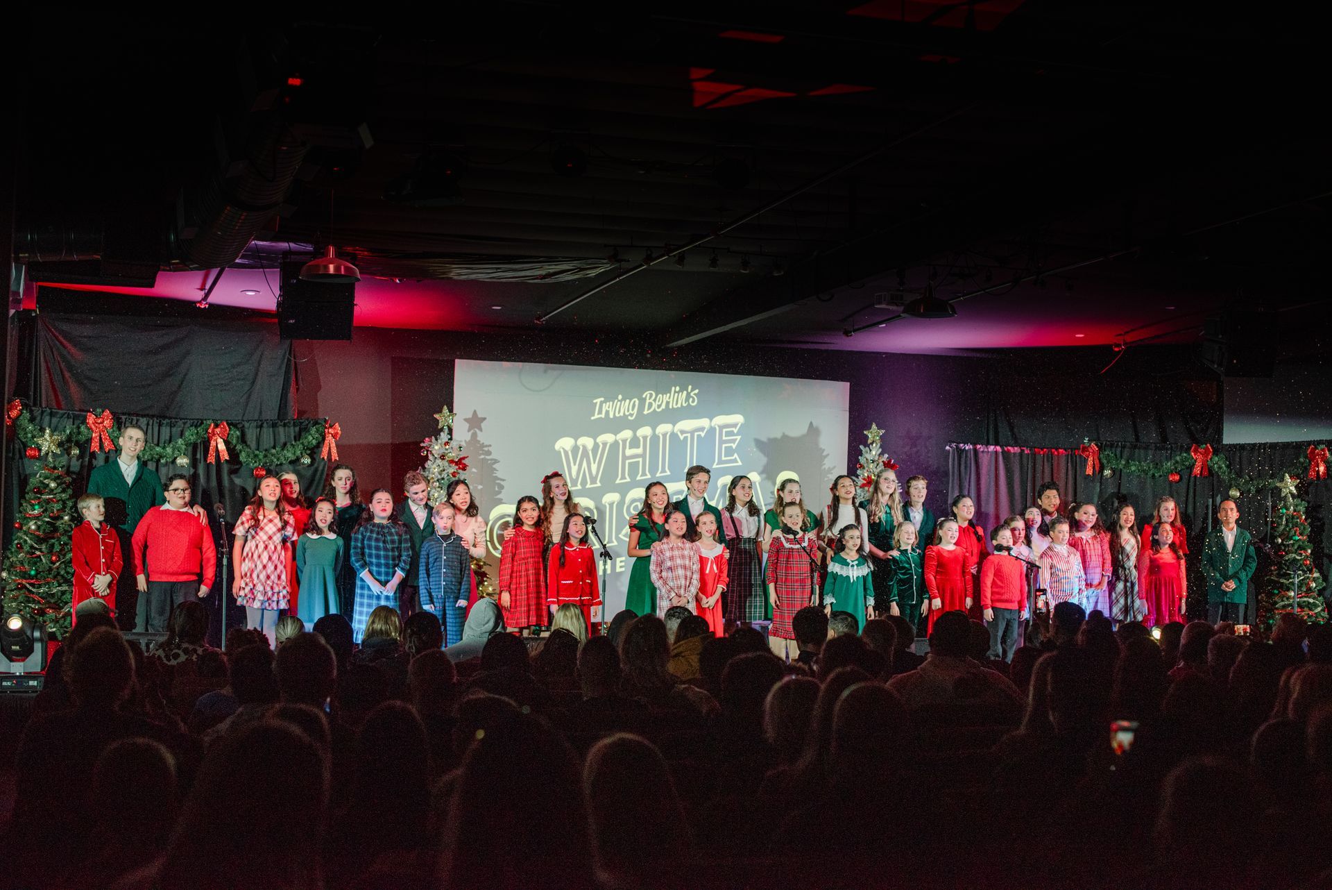 Large choir performing on a stage with pink lighting and a projection screen in the background