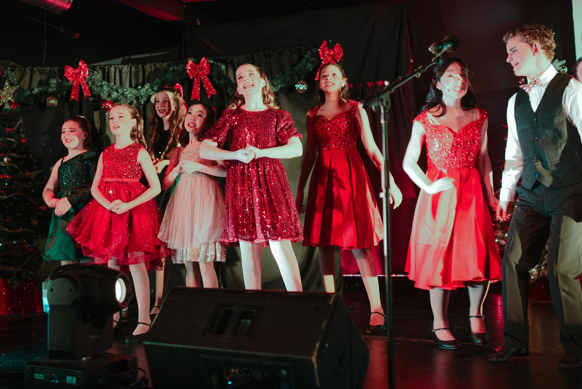 Holiday choir singing on stage in red outfits, holding microphones under festive lights