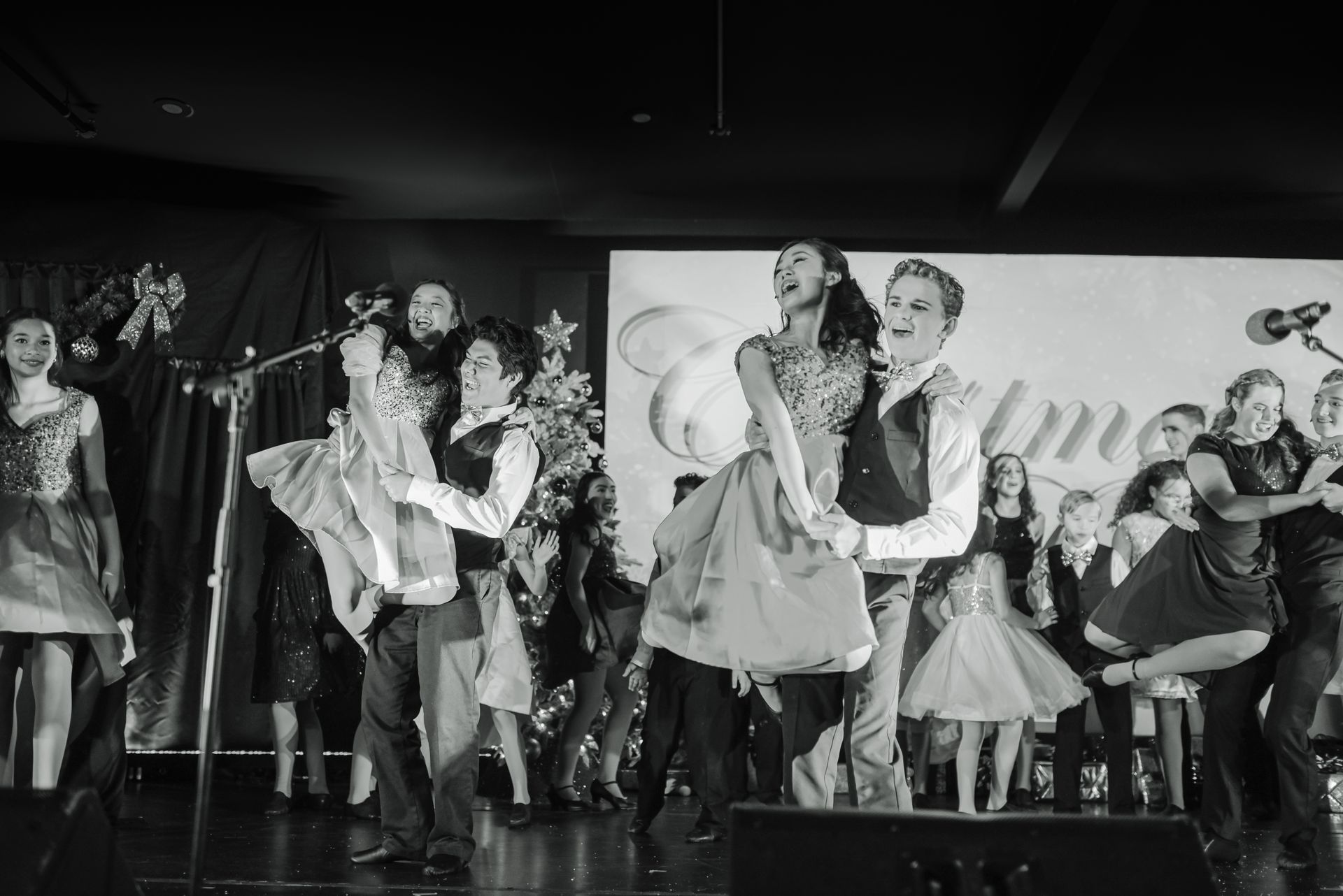 Dancers performing onstage in a lively group routine, with a banner backdrop and audience seated behind them