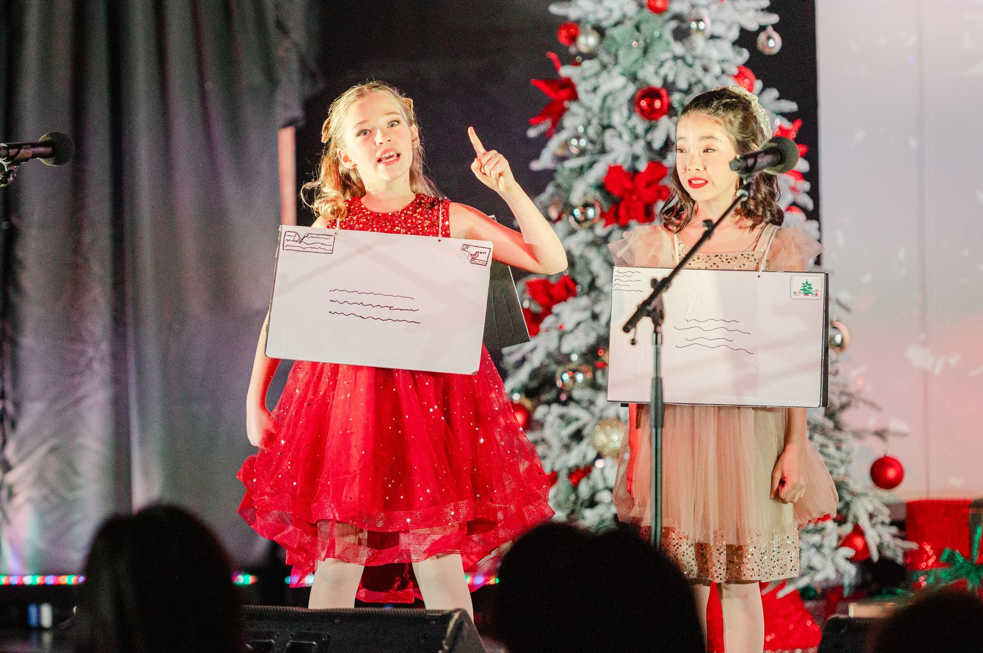 Two people in festive dresses singing onstage with microphones beside a Christmas tree.