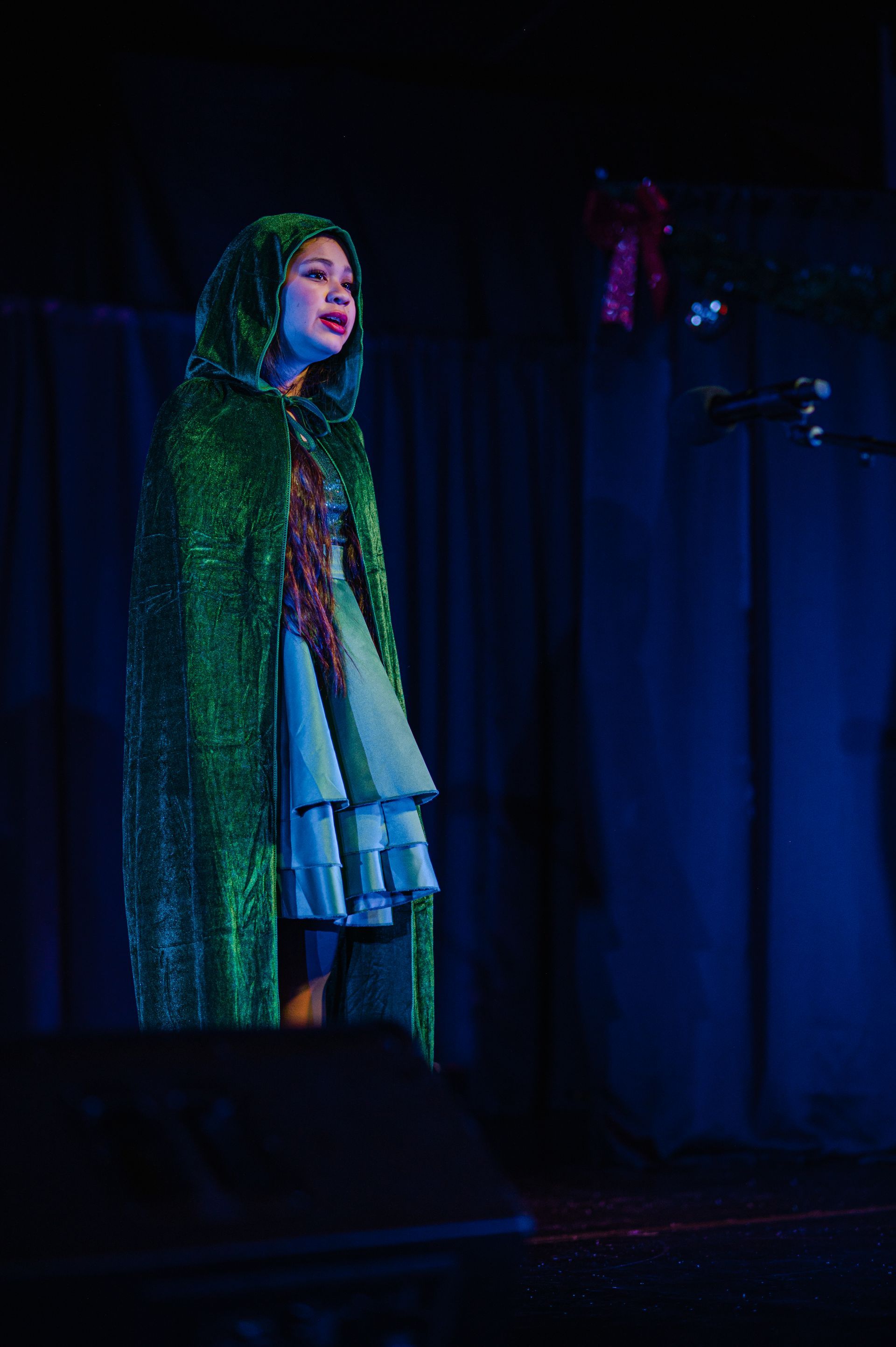 Singer in a shimmering green cape performing on a dimly lit stage under blue-purple lights