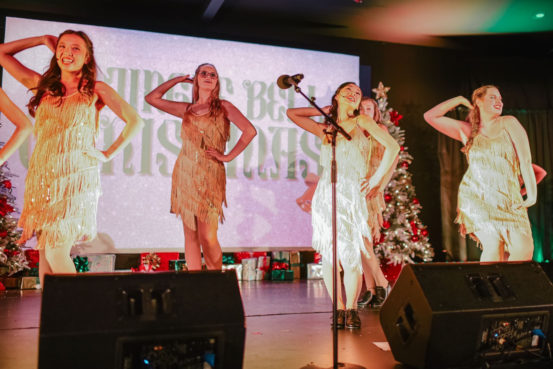 Four performers in gold dresses pose on stage in front of microphones and a lit holiday backdrop.