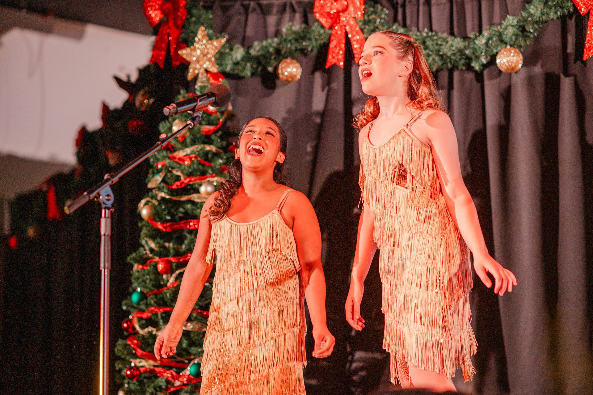 Two singers in gold dresses performing on a decorated holiday stage with a Christmas tree