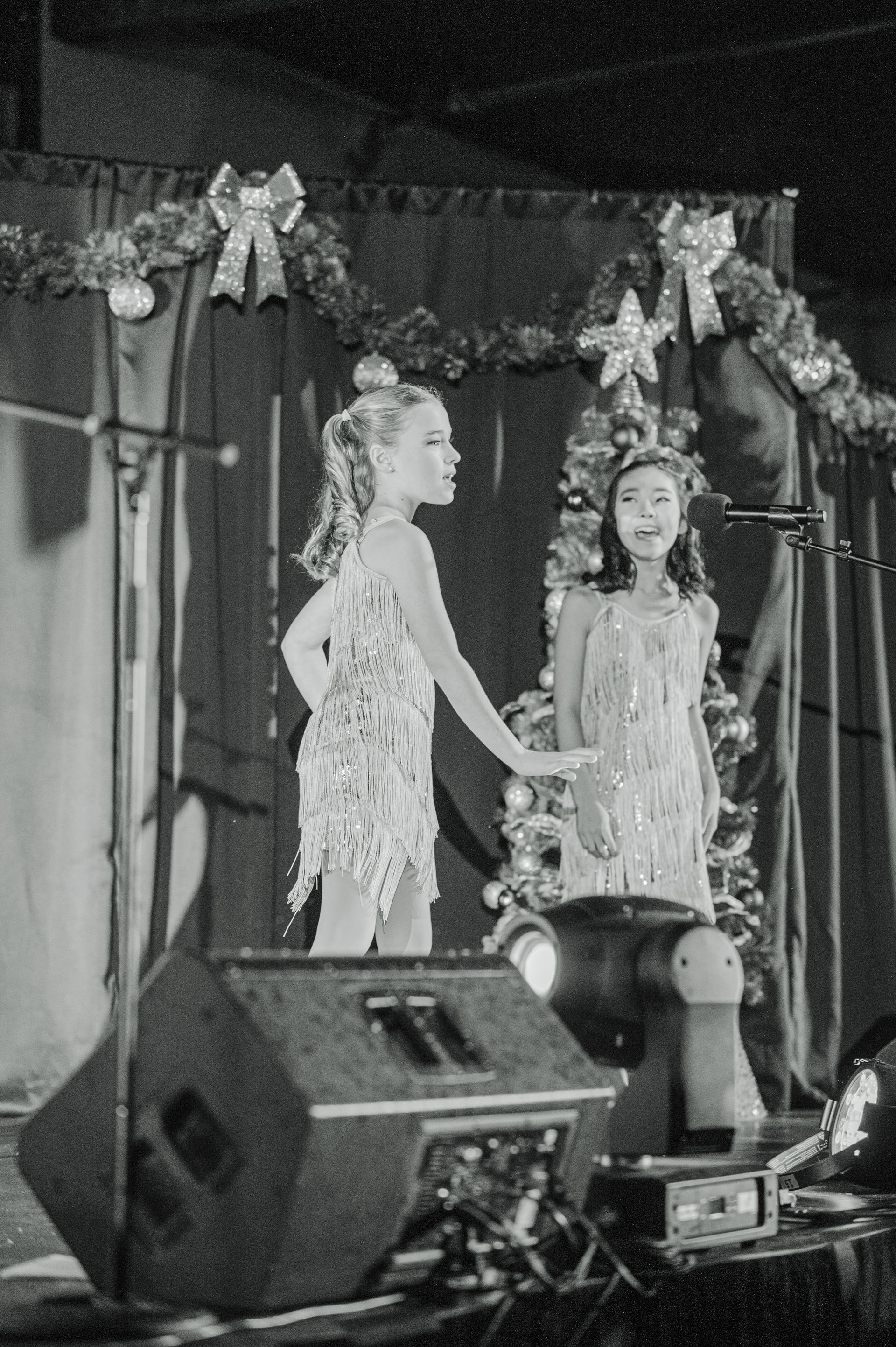 Two performers in feathered costumes stand on a decorated stage beside microphones and a speaker.