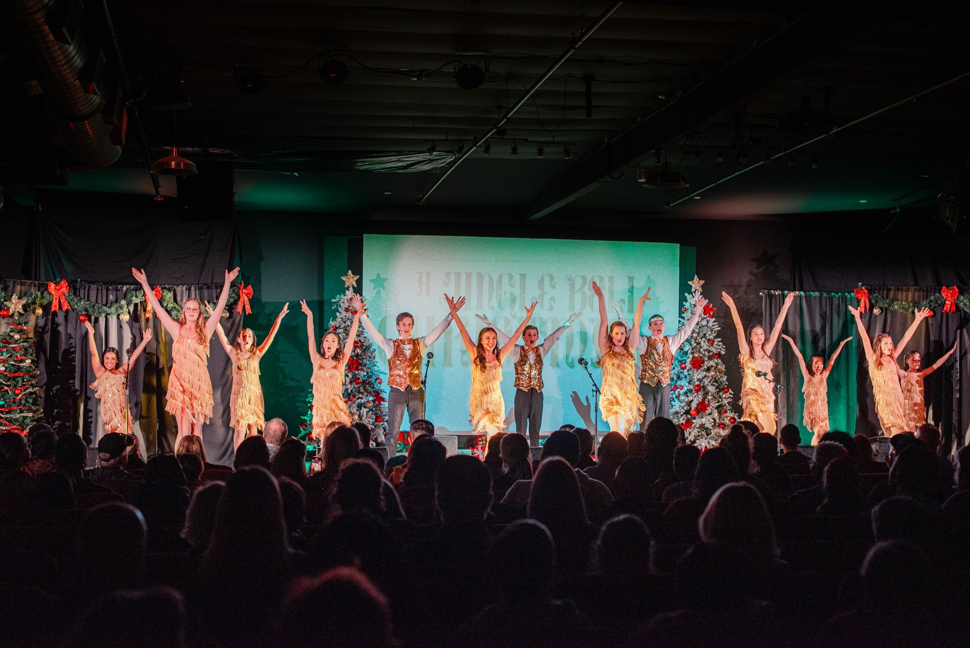 Performers in colorful costumes on a stage with raised arms, lit by green and warm lights, facing an audience