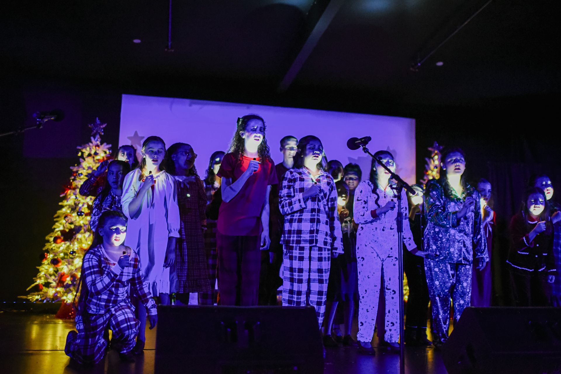 Choir singing on a dark stage under purple-blue lights, with a lit Christmas tree at left