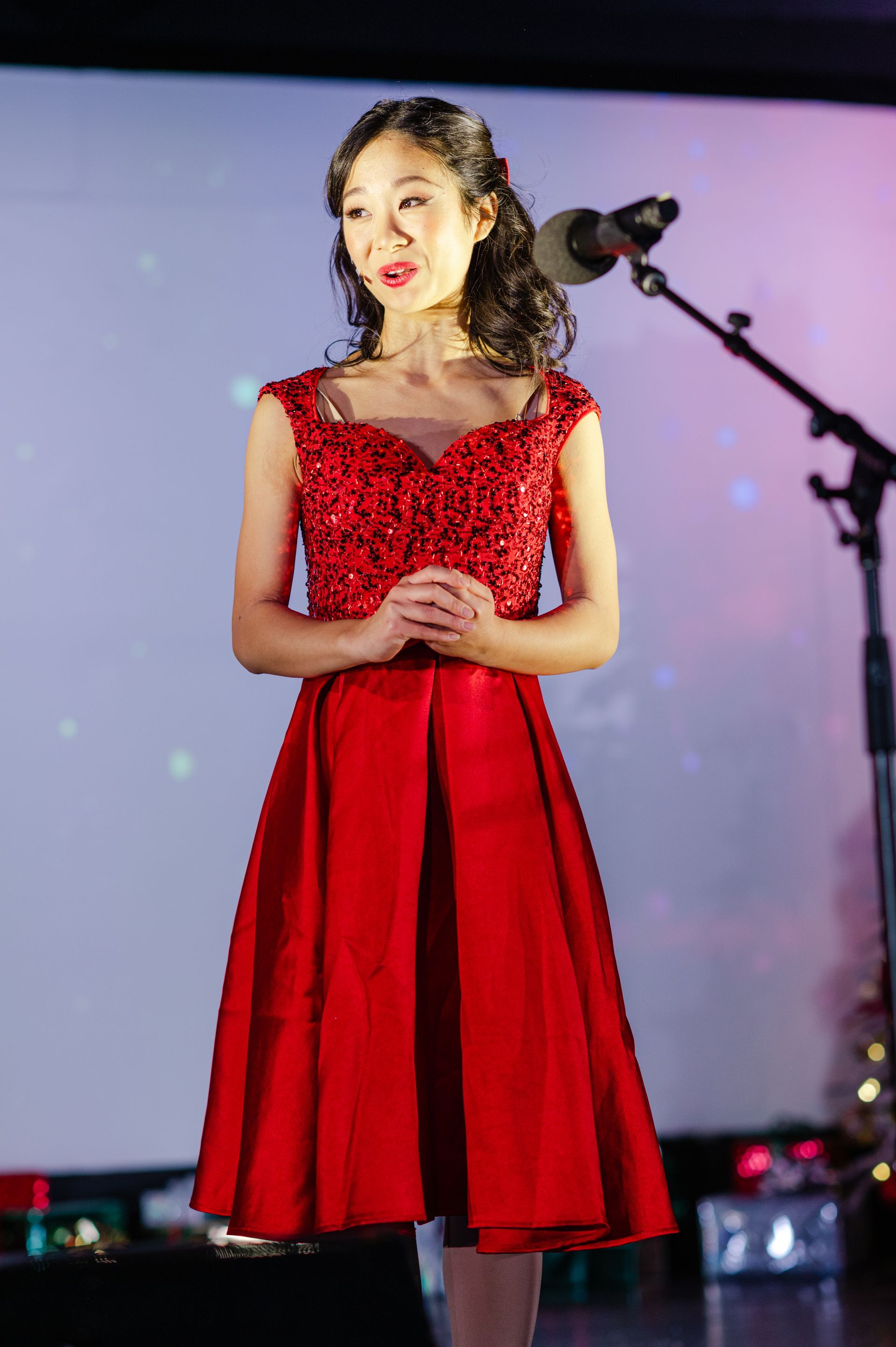 Singer in a red dress onstage beside a microphone, performing under soft stage lights.
