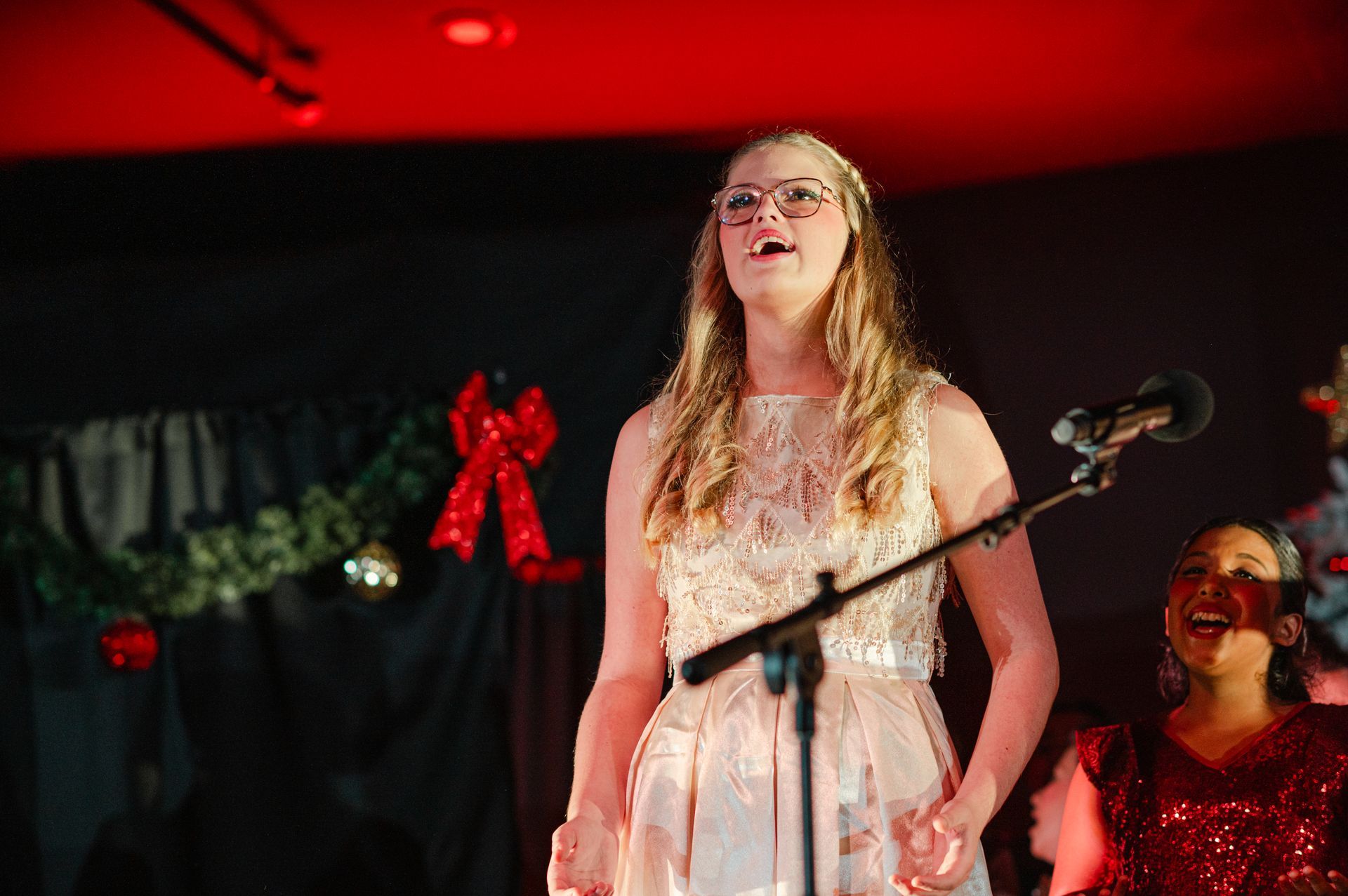 Woman in a white dress singing on a stage under red lights, with a microphone and another performer nearby
