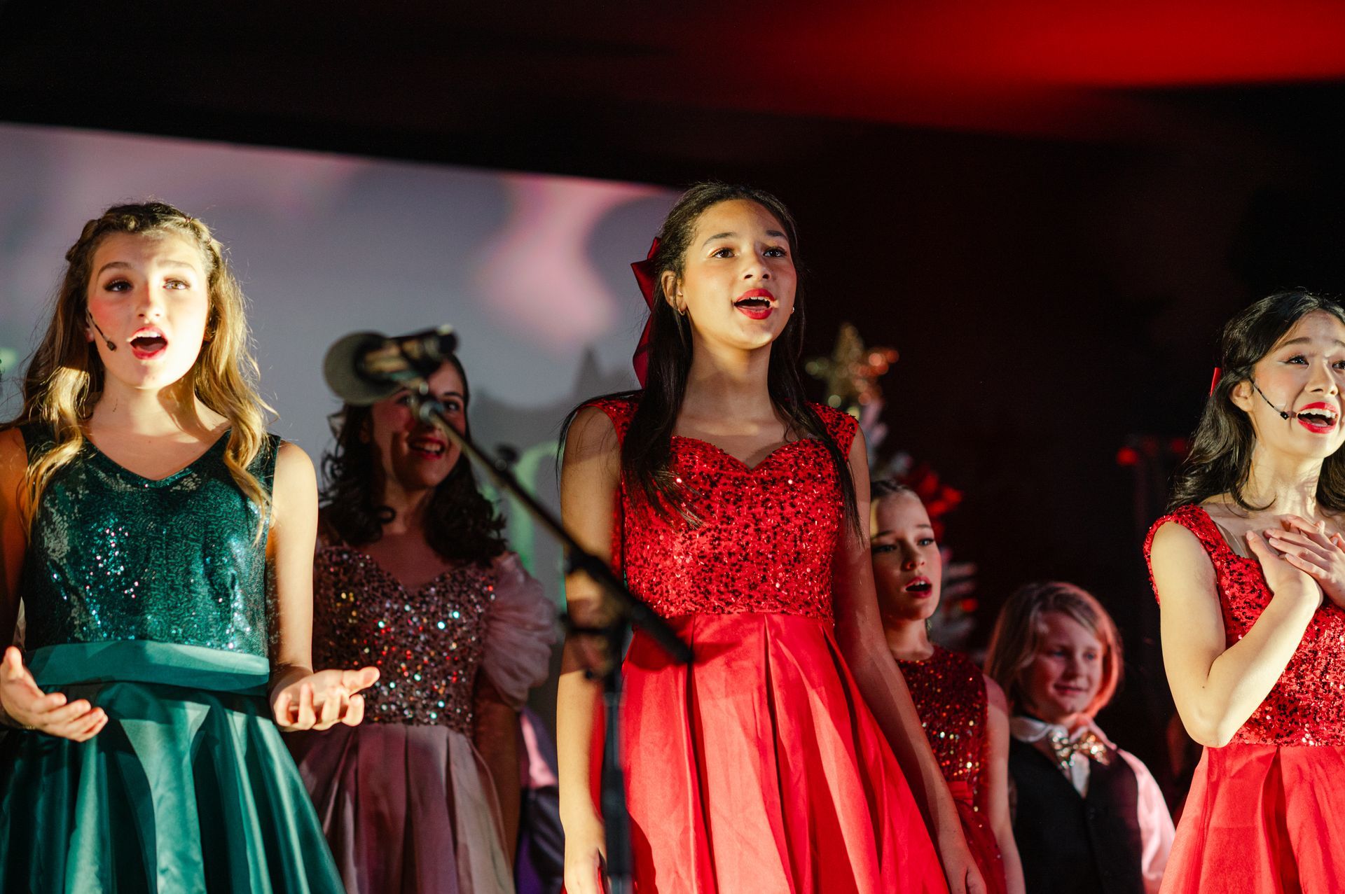 Children performing on stage in red and green dresses, singing under stage lights