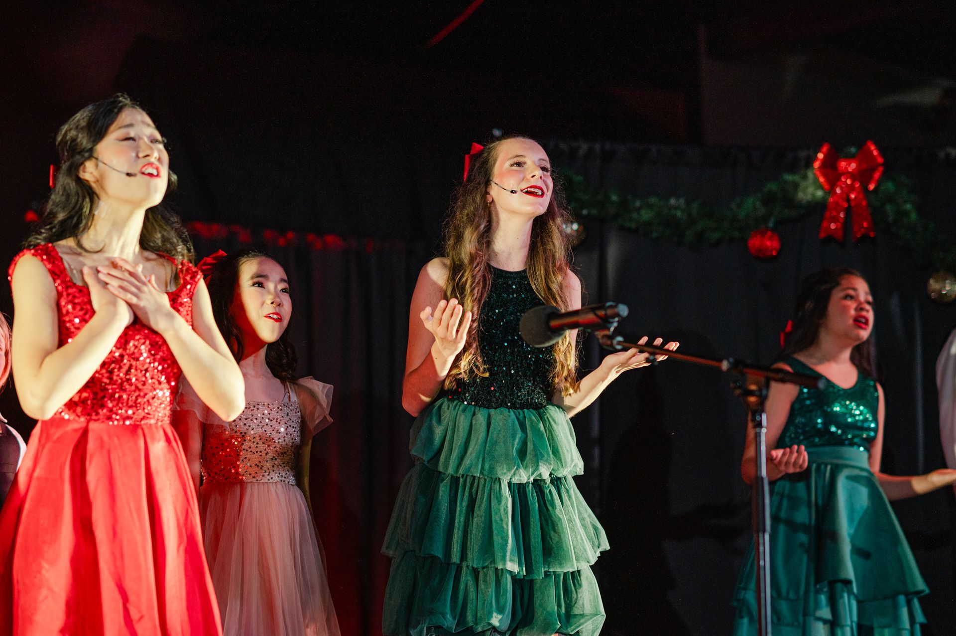 Four performers in sparkling red, pink, green, and teal dresses singing onstage with microphones