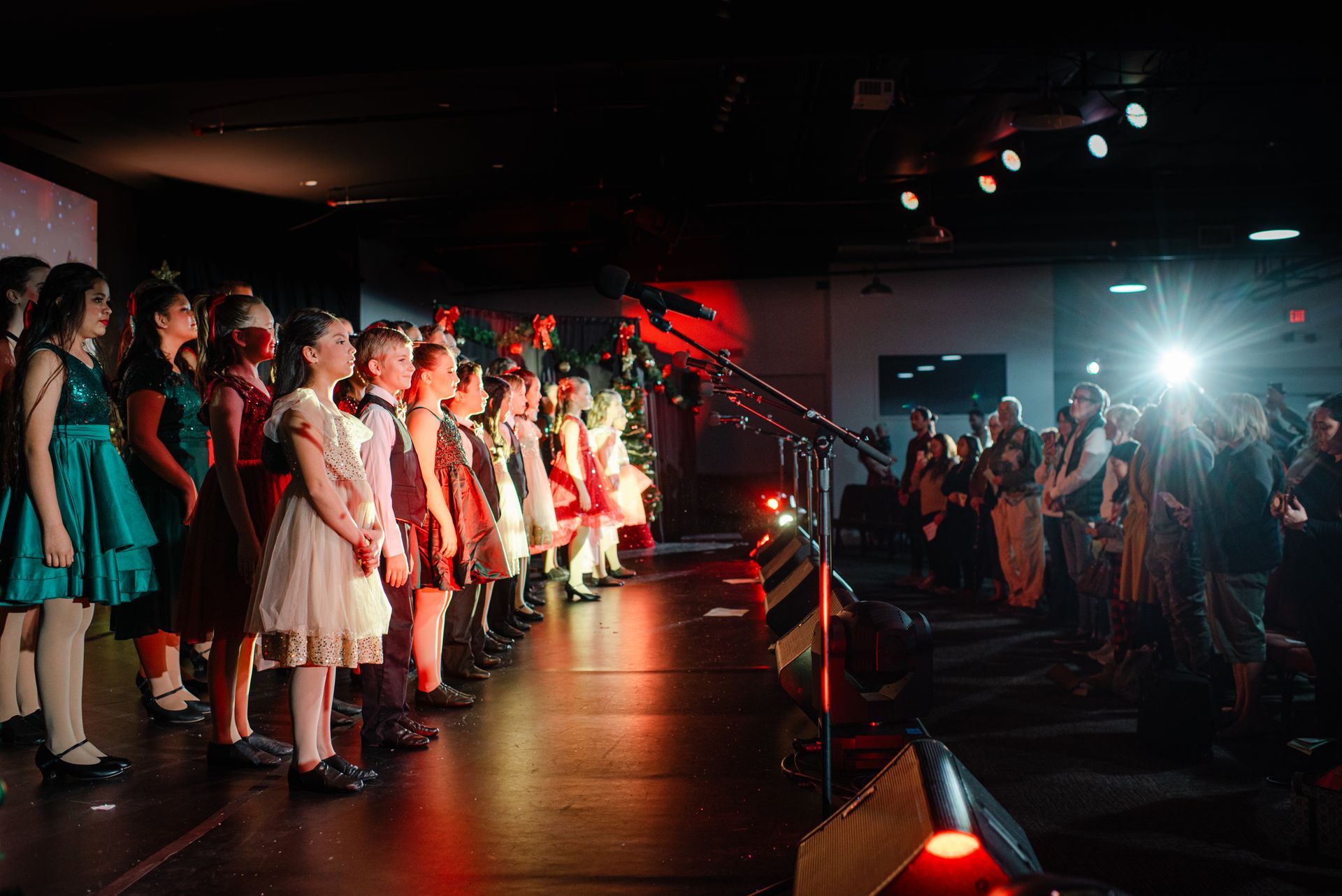 Children singing on a stage under red lights while an audience watches and records the performance