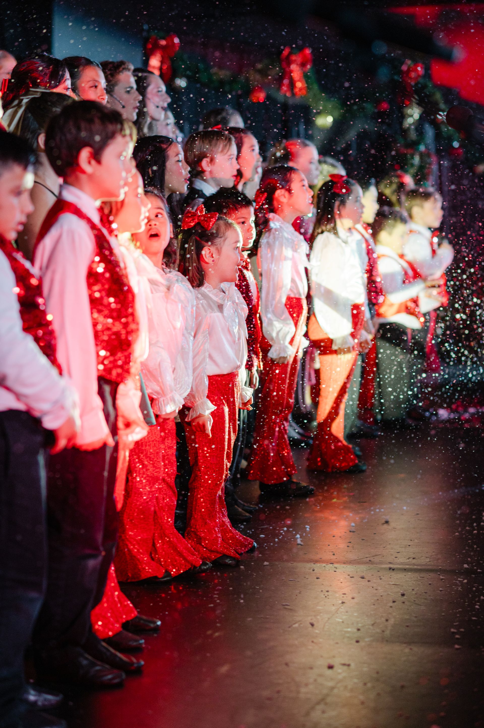 Children in white shirts and red pants standing in a row amid falling confetti on a stage