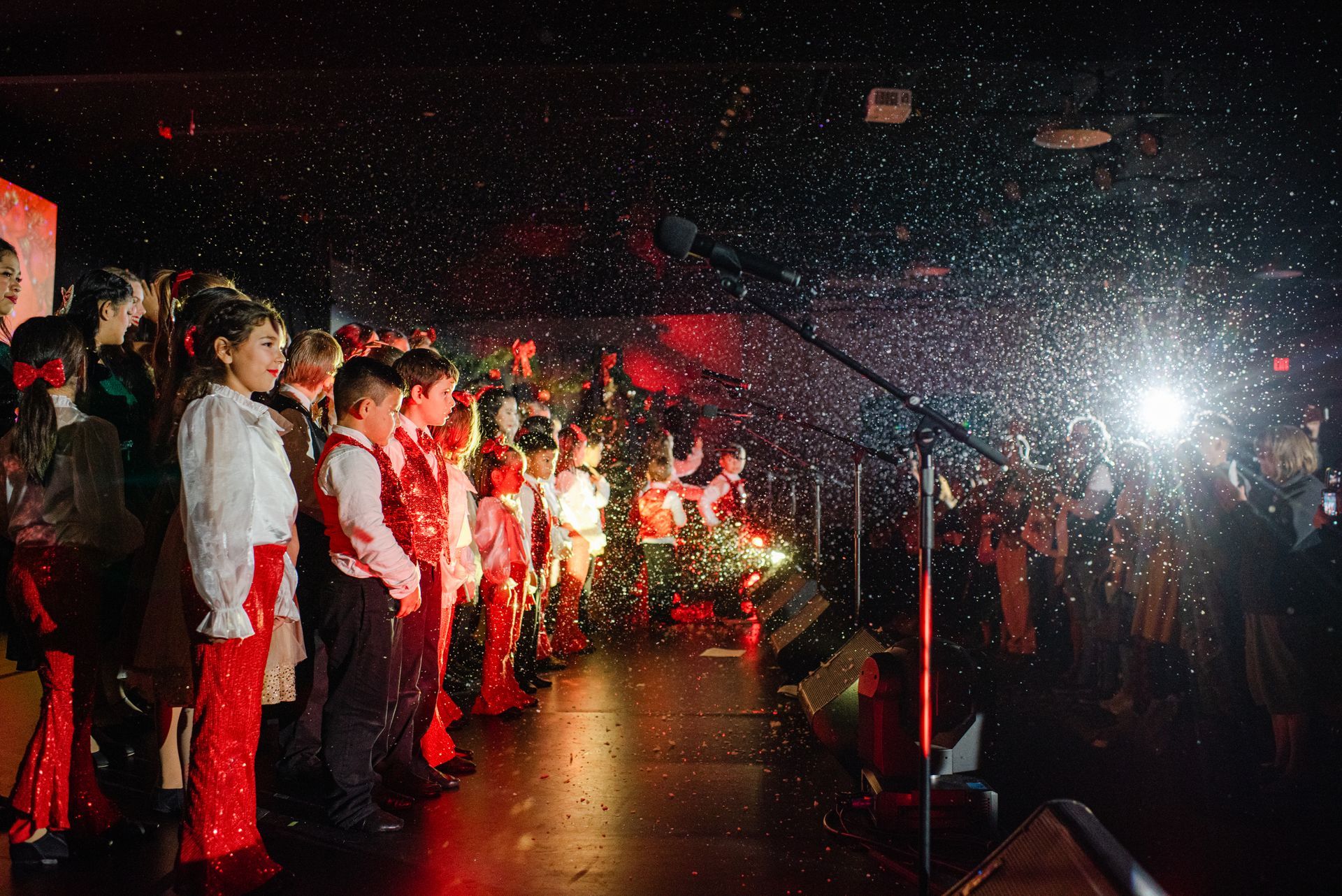Children singing on a dark stage with red lighting and a bright camera flash in the audience