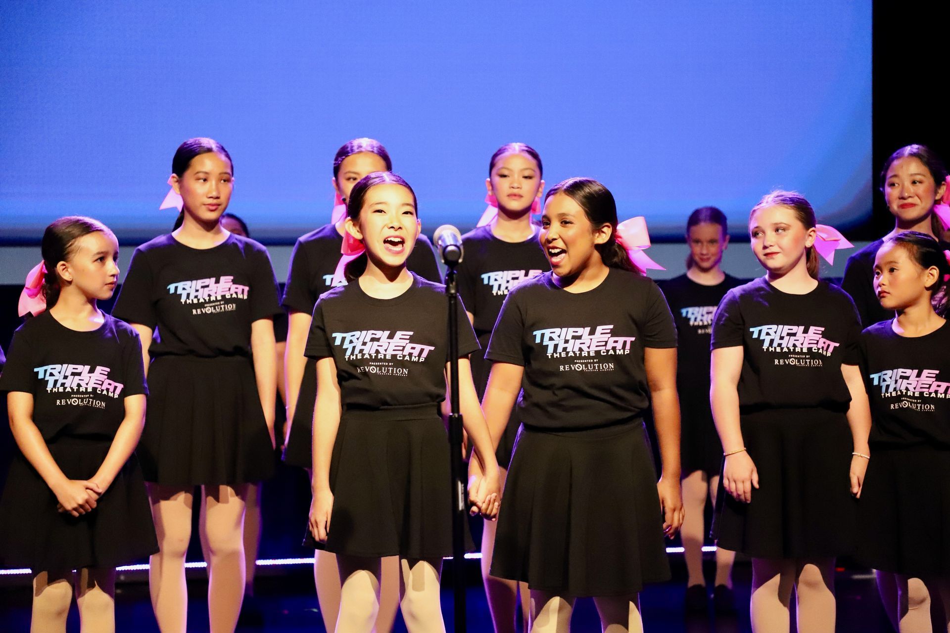 Group of dancers in black T-shirts standing onstage under blue lighting for a theater showcase.