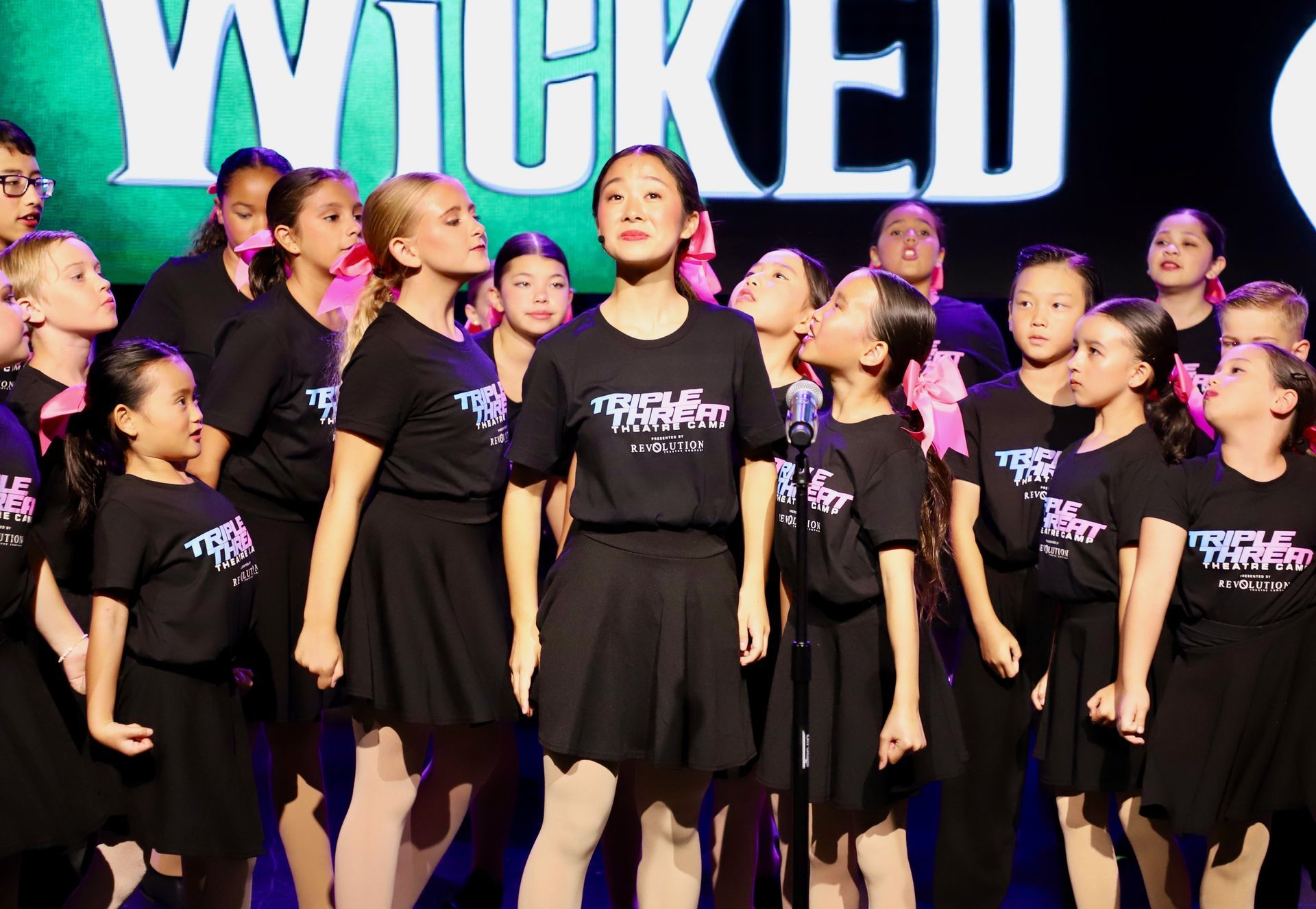 Children in black outfits performing on a stage with a large “WICKED” backdrop for a theater showcase.
