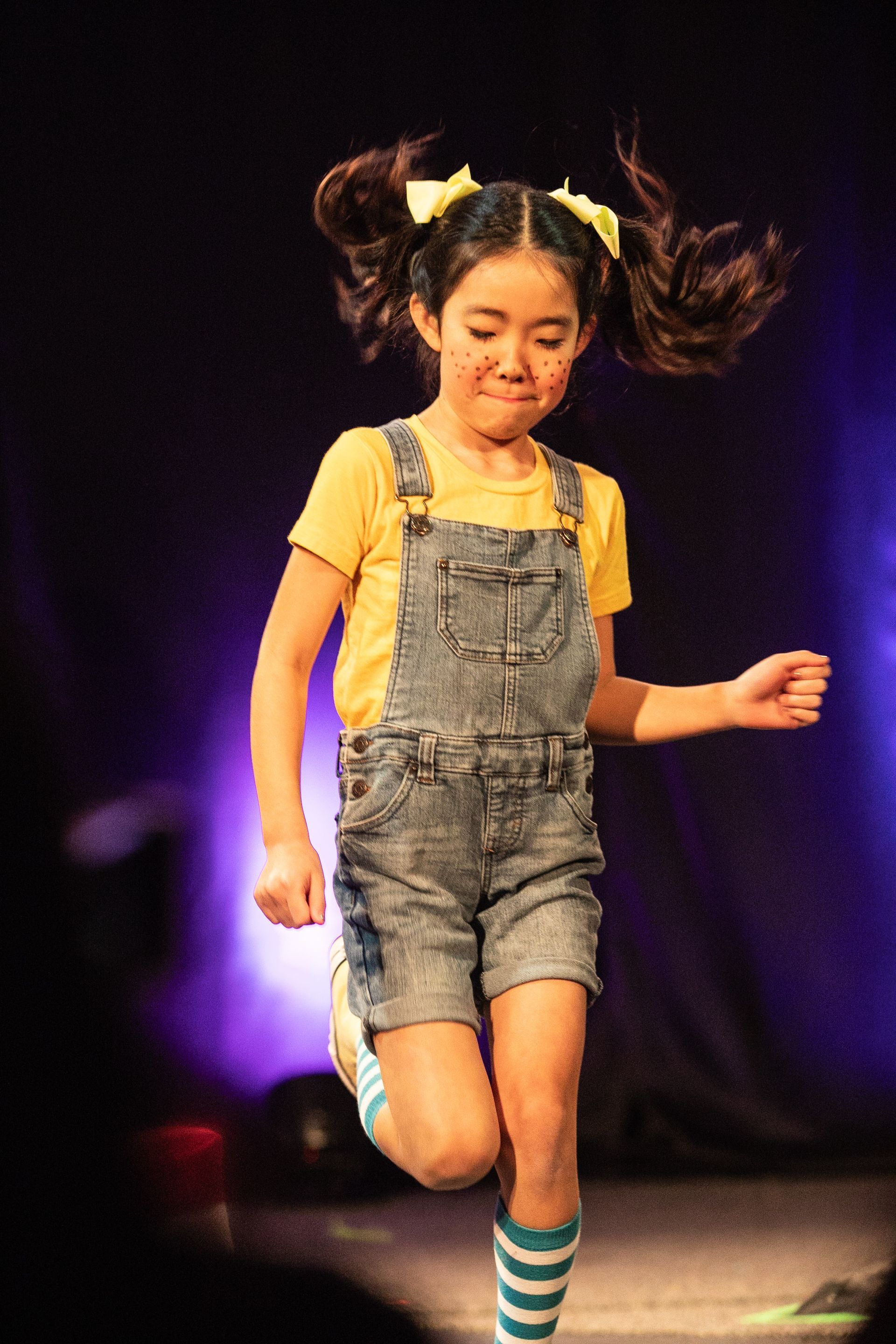 A little girl wearing overalls and striped socks is dancing on a stage.