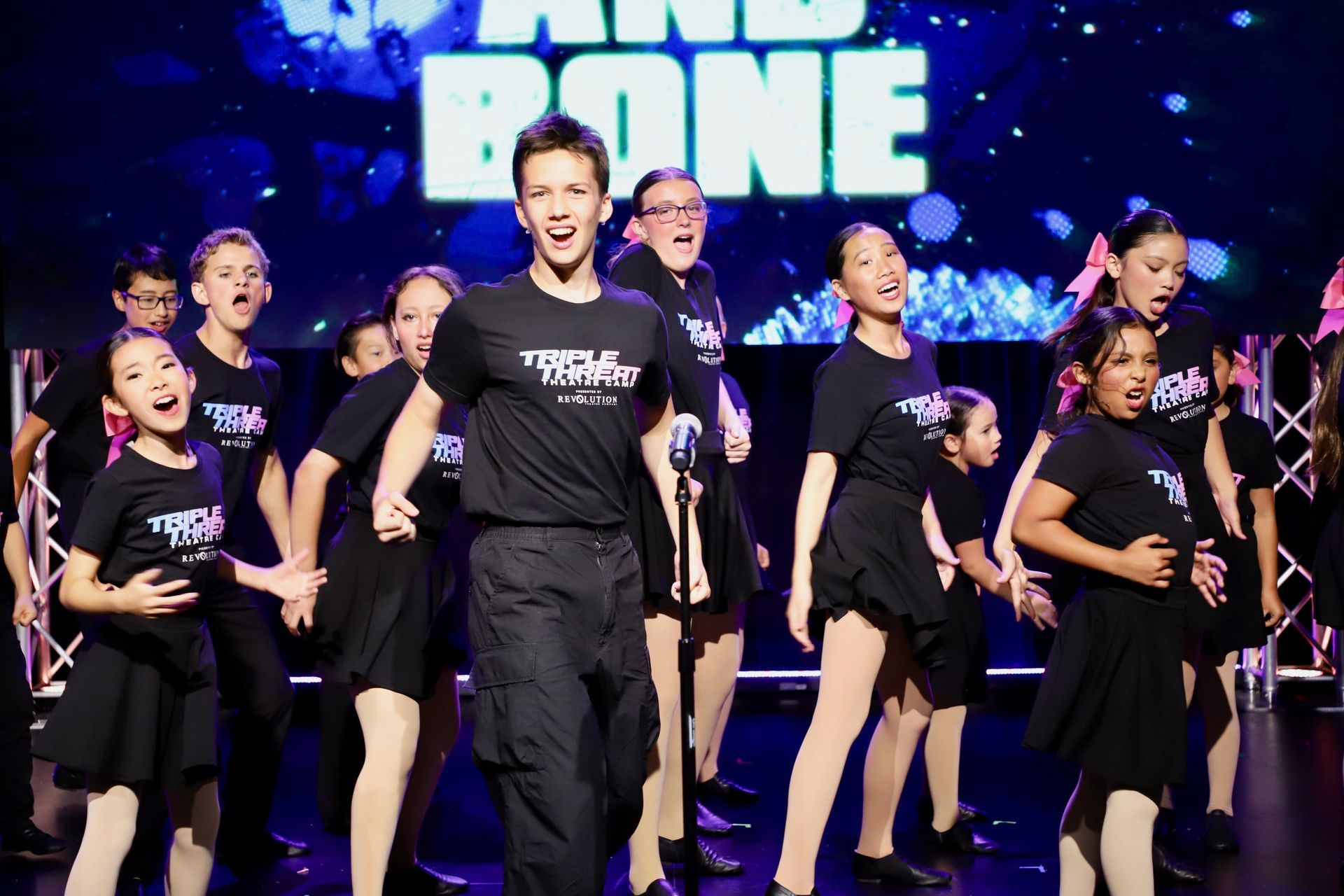 A group of performers in black shirts dance onstage under blue lights, with one man centered in front for a theater showcase.