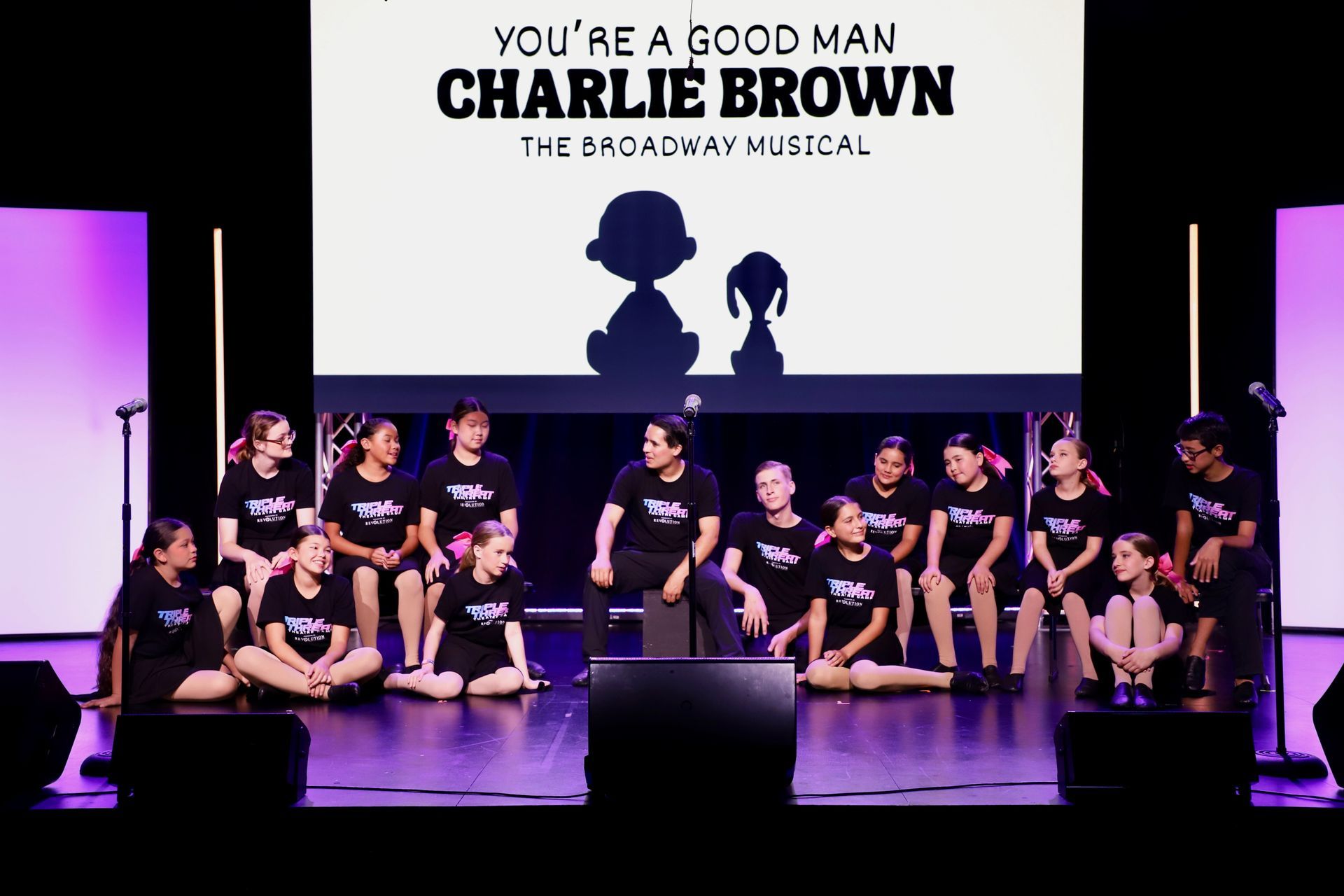 Cast of “You’re a Good Man, Charlie Brown” seated on a theater stage under purple lighting for a theater showcase.