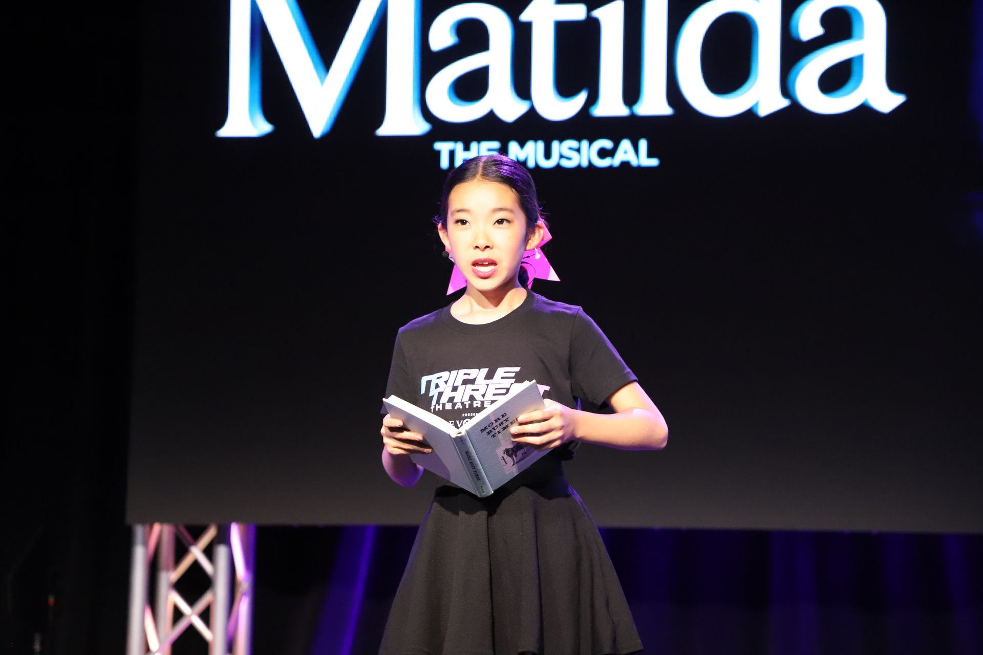 Performer on stage holding a script in front of a Matilda the Musical backdrop for a theater showcase.