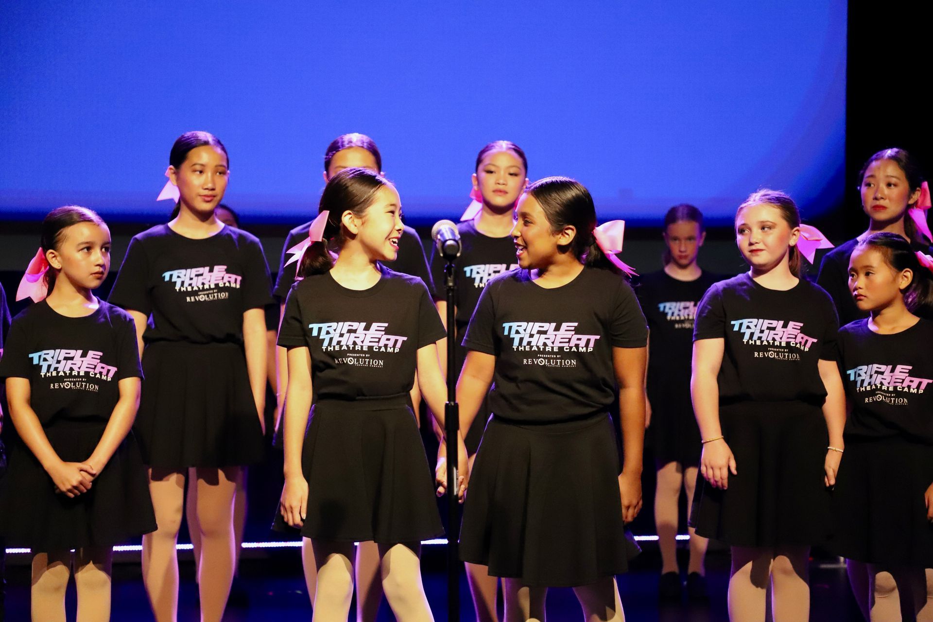 A group of dancers in black shirts stand onstage against a blue-lit backdrop for a theater showcase.