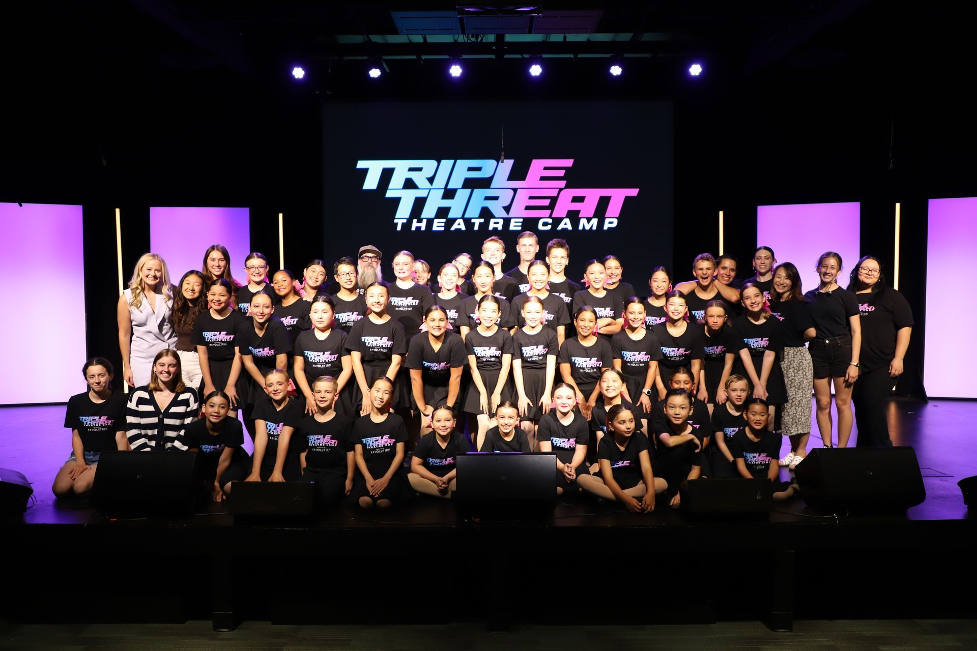 Large group of dancers posing on a stage with purple lights and a “Triple Threat” backdrop for a theater showcase.