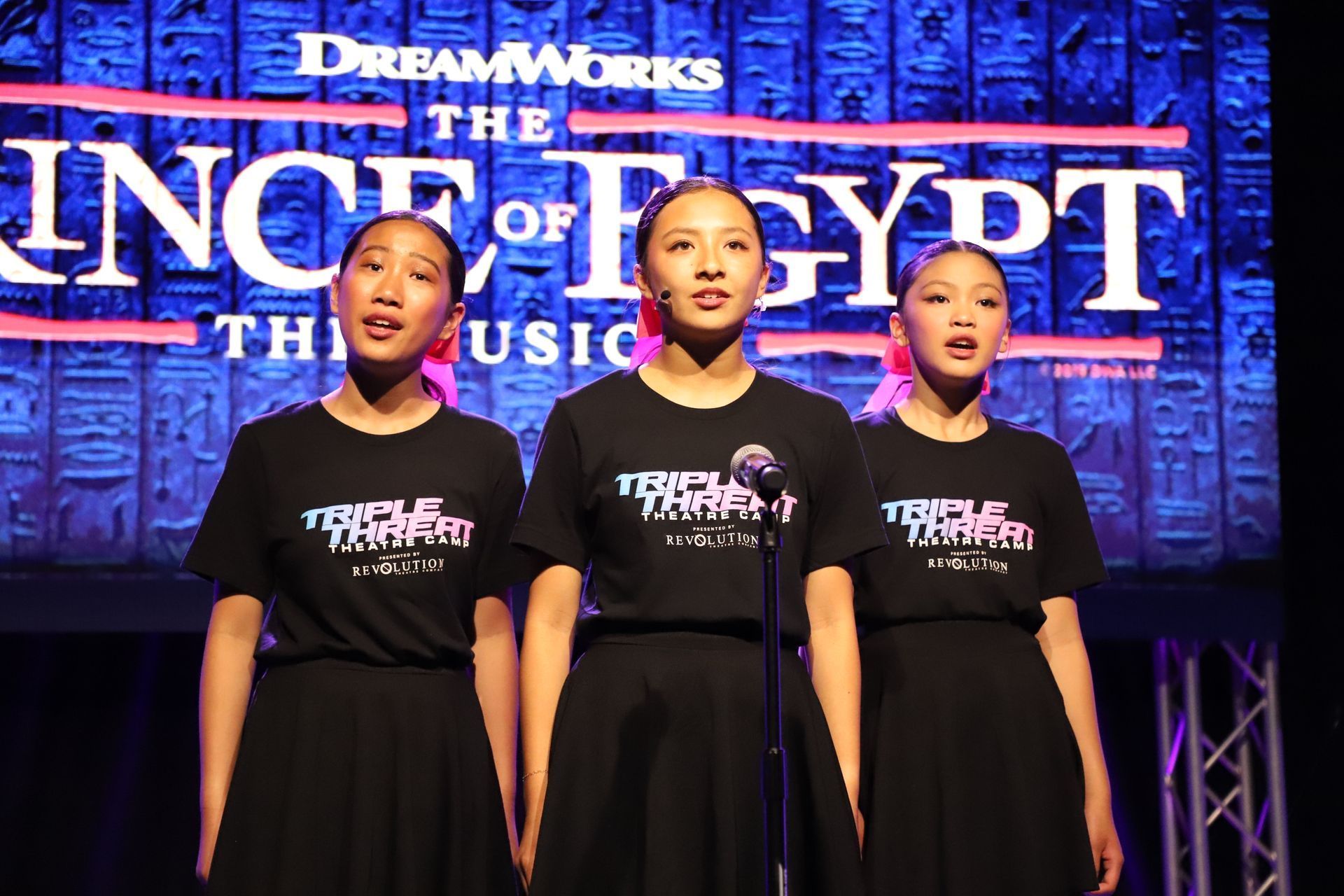 Three singers in black dresses perform onstage before a “Prince of Egypt” backdrop for a theater showcase.