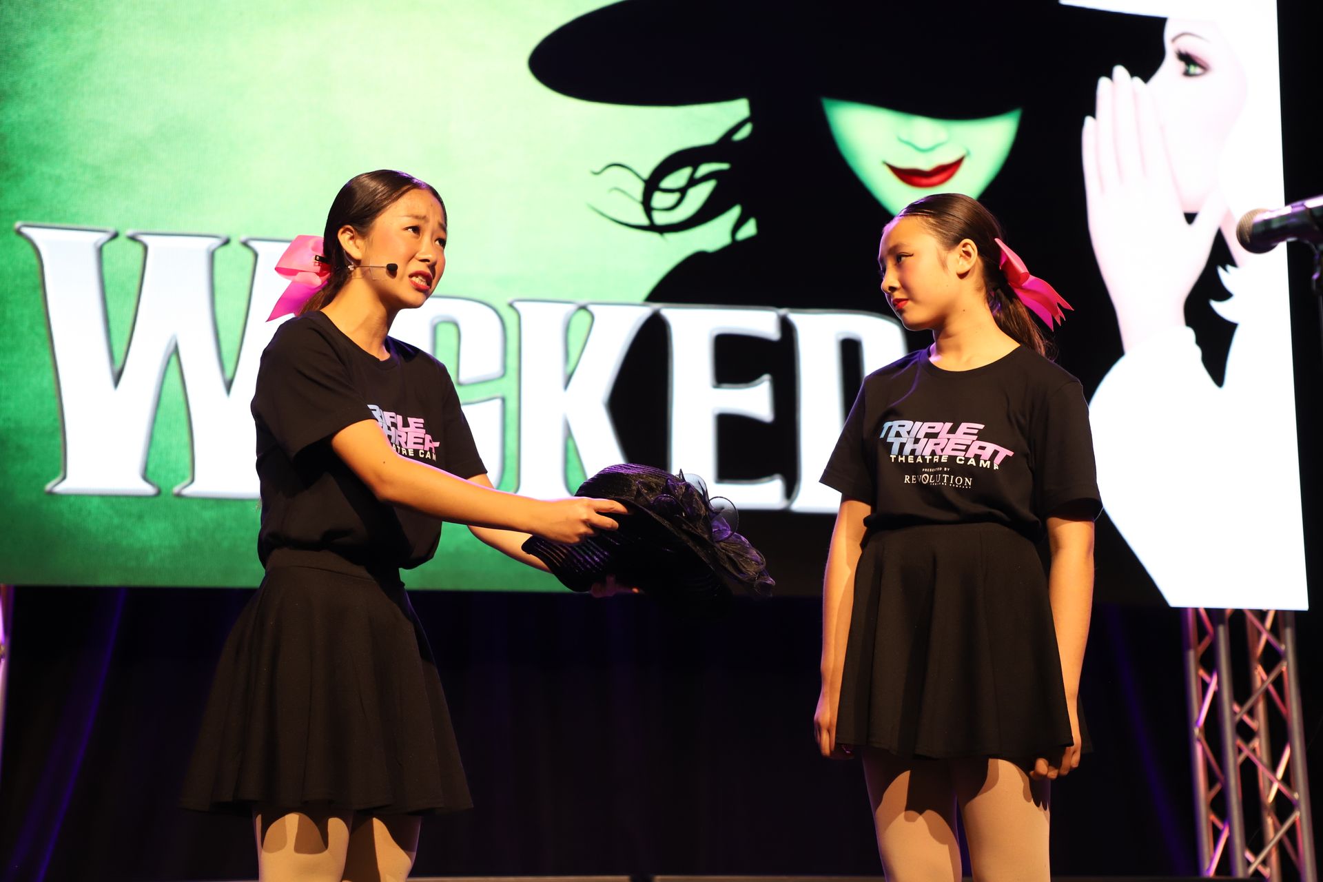 Two women in black dresses on a stage, facing each other before a Wicked-themed backdrop for a theater showcase.