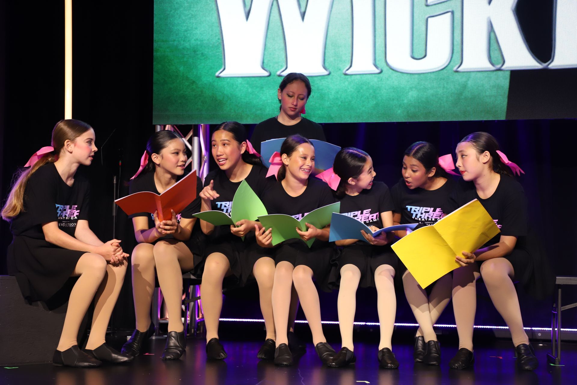 Children seated onstage, singing from colorful songbooks in a group performance