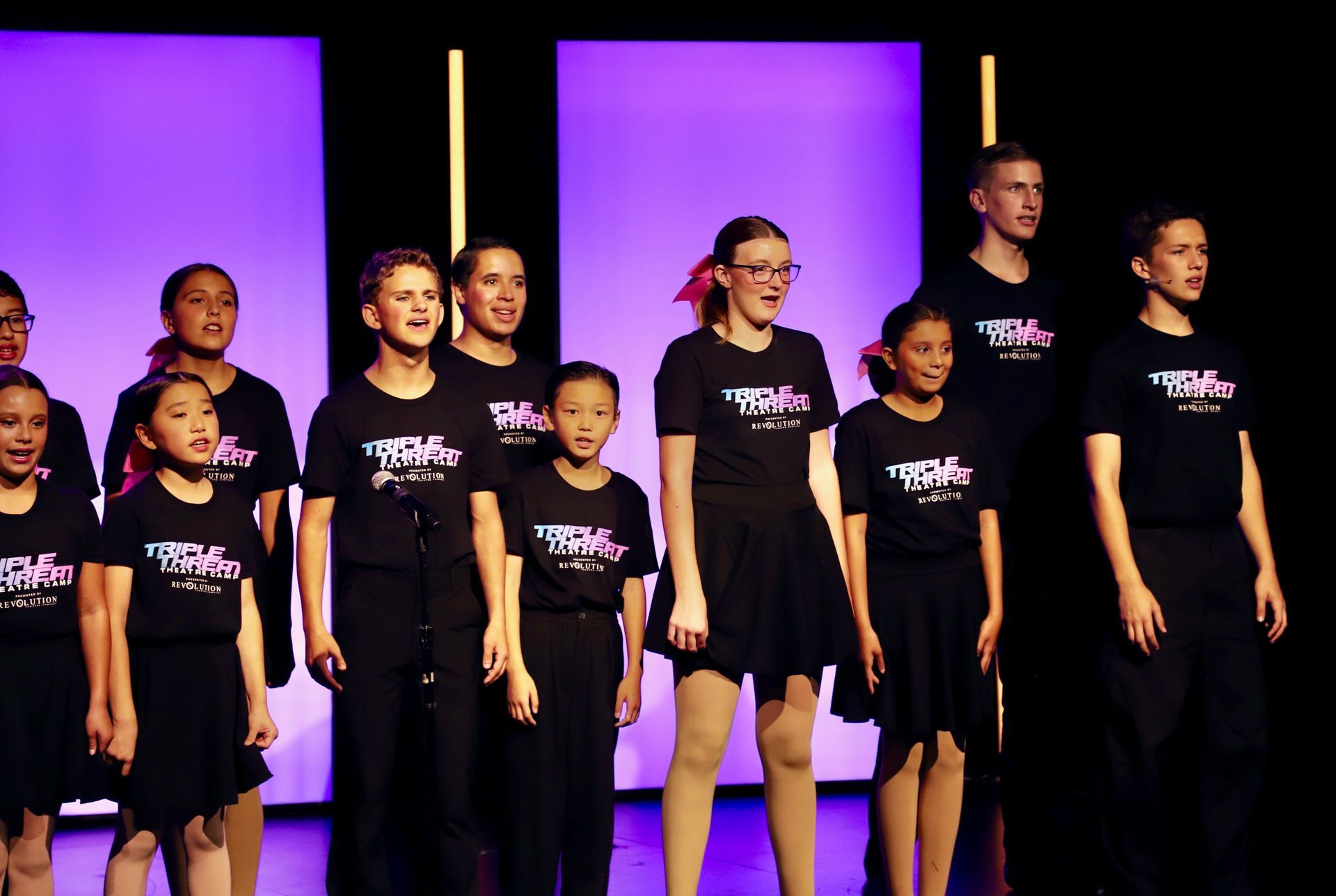 Children and adults in black T-shirts standing on a stage with purple-lit panels for a theater showcase.