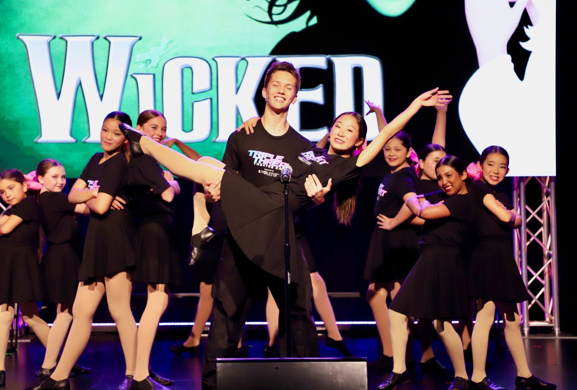 Dance troupe performing onstage in black outfits before a Wicked backdrop for a theater showcase.