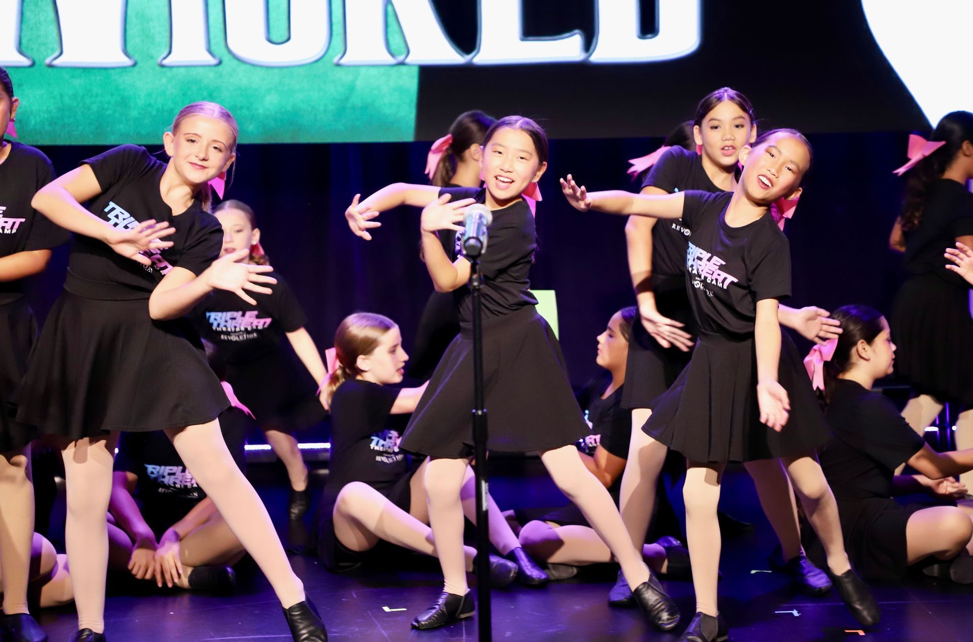 Group of dancers in black outfits performing a choreographed routine onstage under purple lighting for a theater showcase.