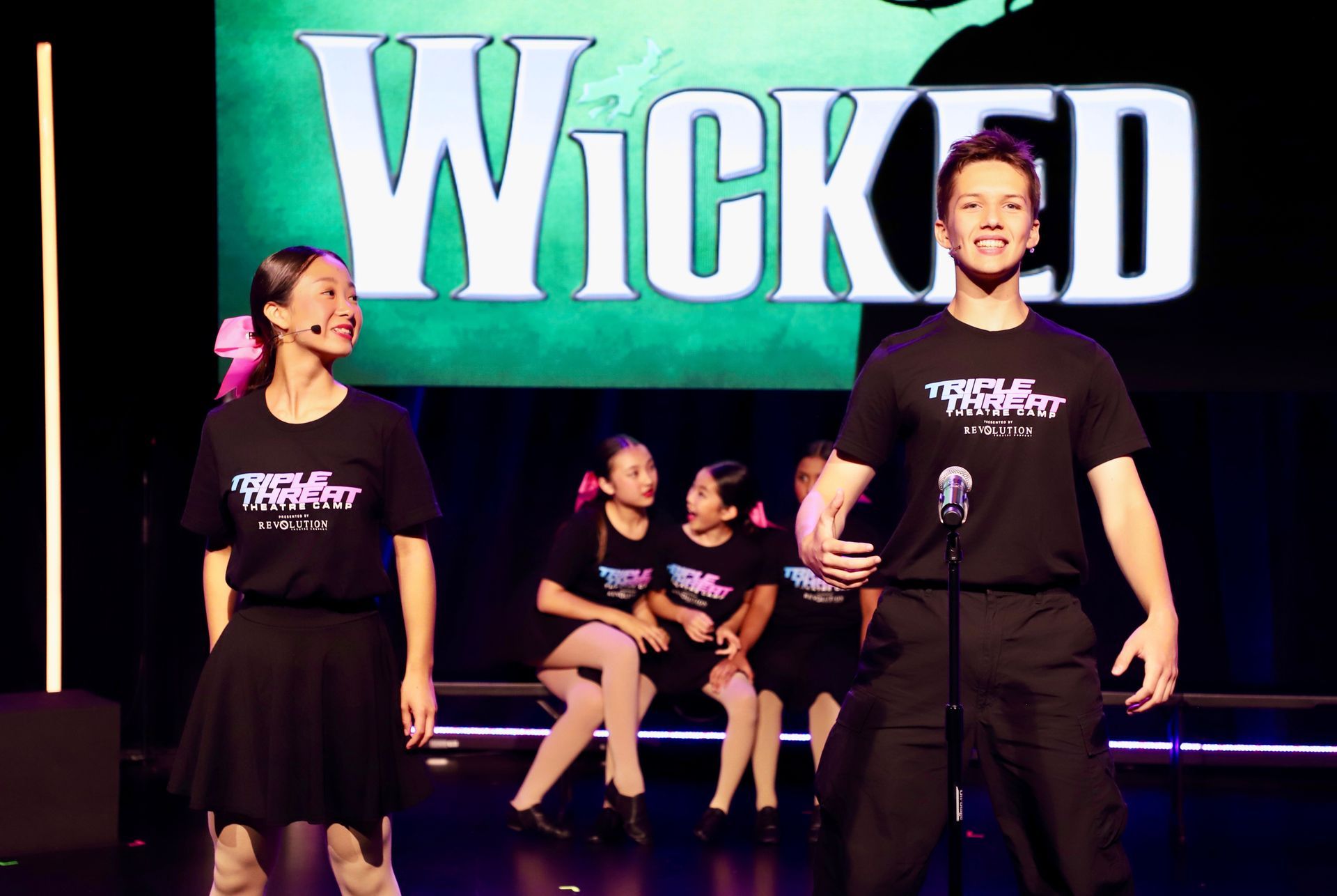 Two performers in black shirts on stage with a large Wicked sign behind them for a theater showcase.