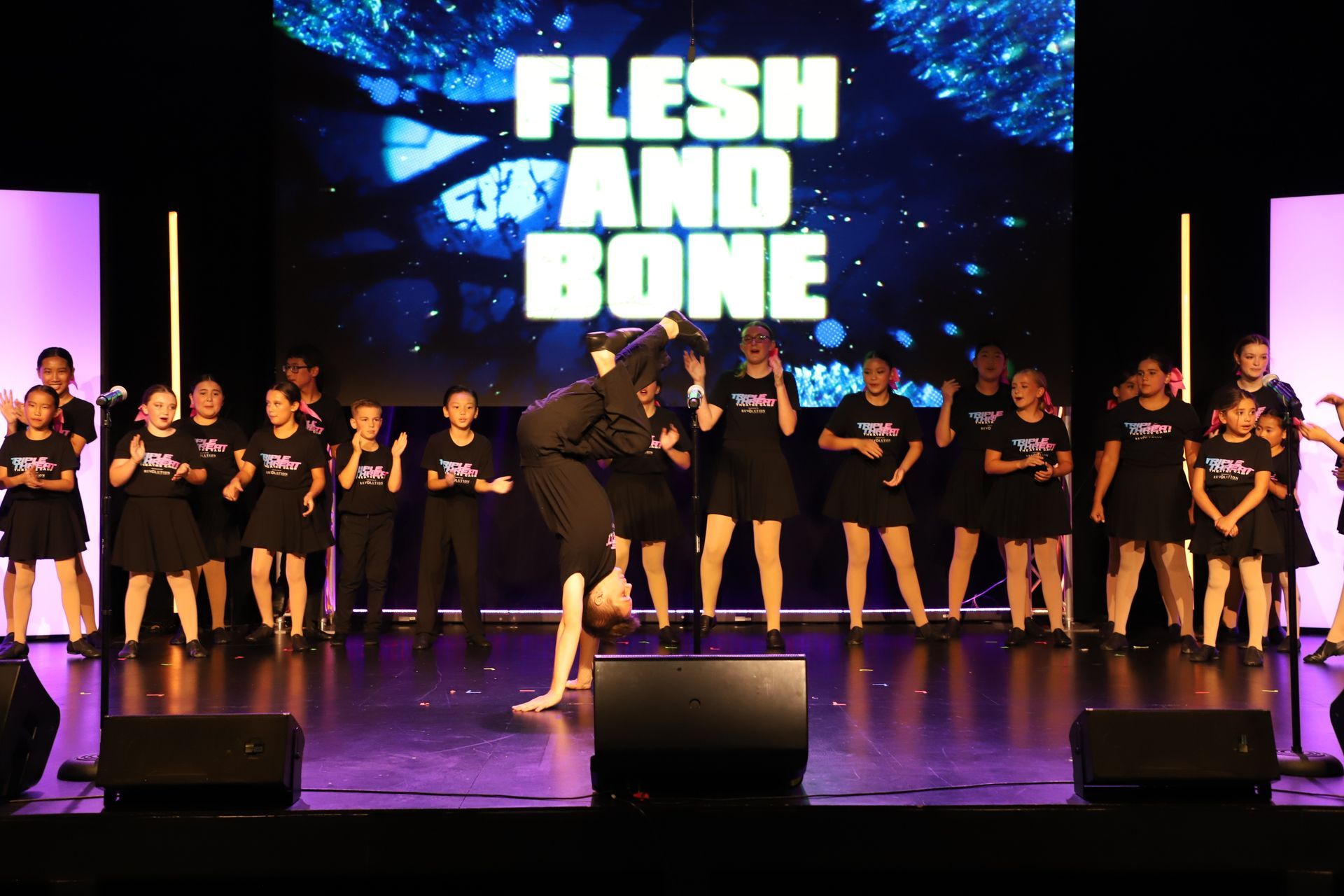 Group of performers onstage in black outfits under a “FLESH AND BONE” screen backdrop for a theater showcase.