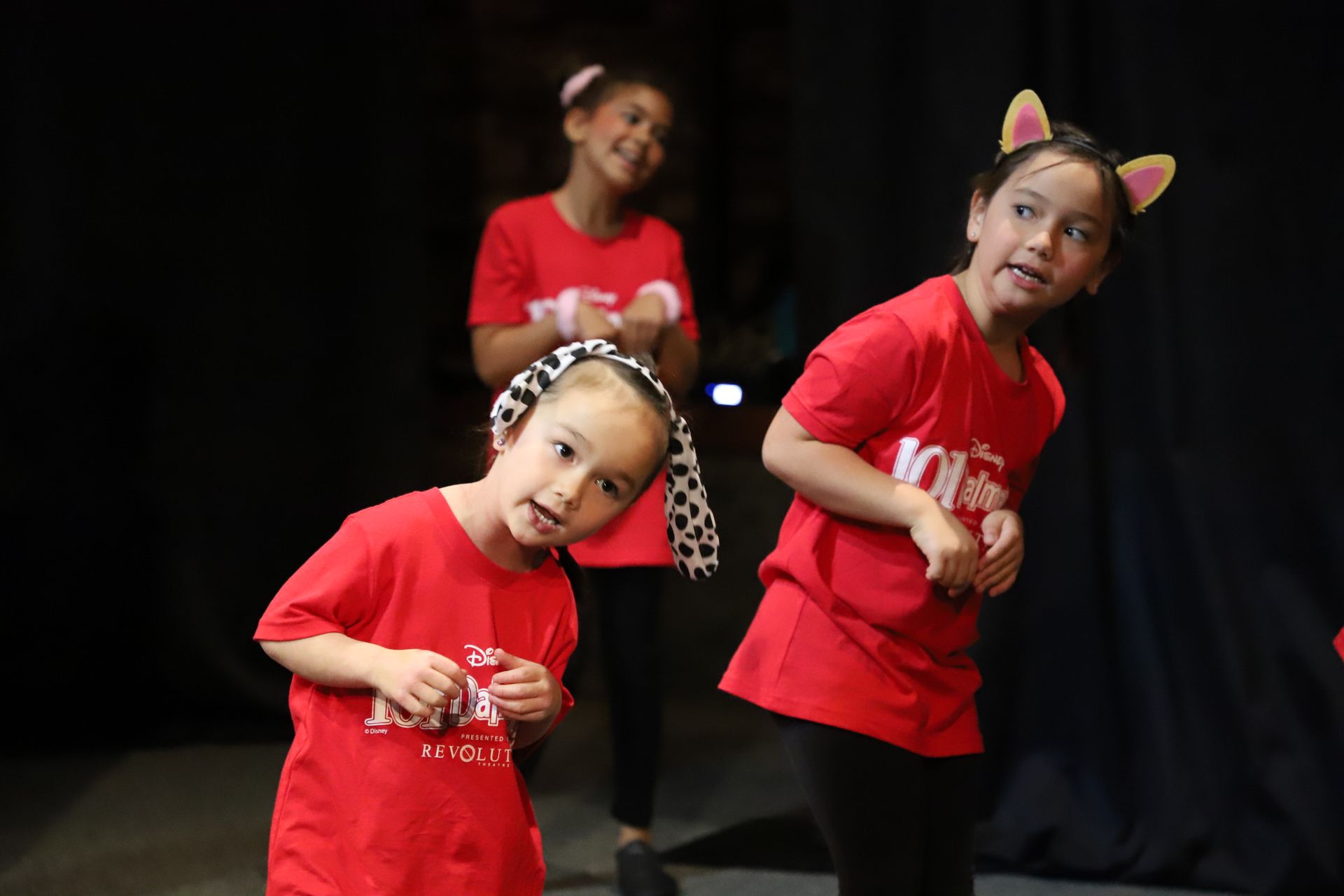 Three little girls in red shirts are dancing on a stage.