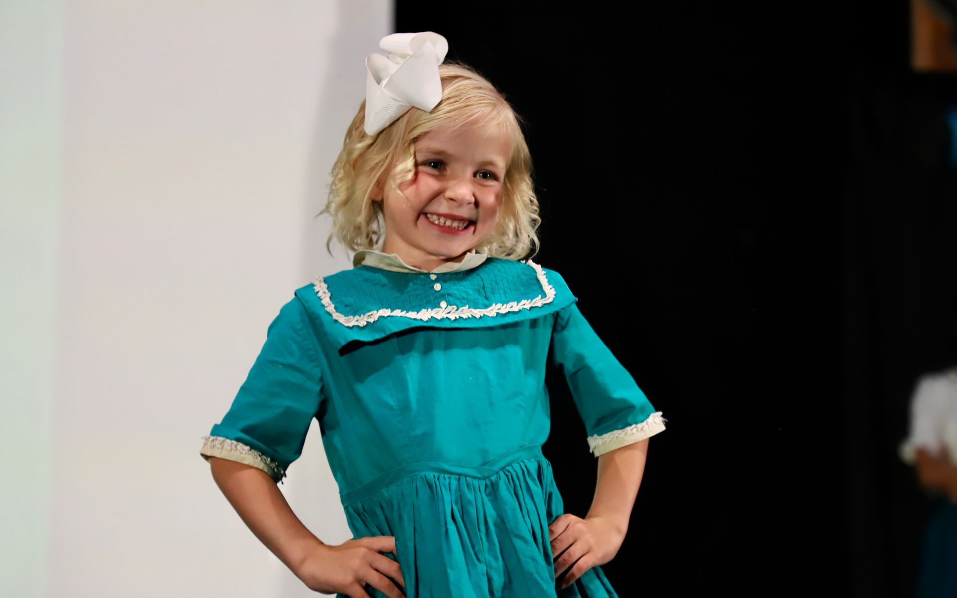 Child in a turquoise dress with white trim and a large white bow, smiling with hands on for a theater performance.hips.