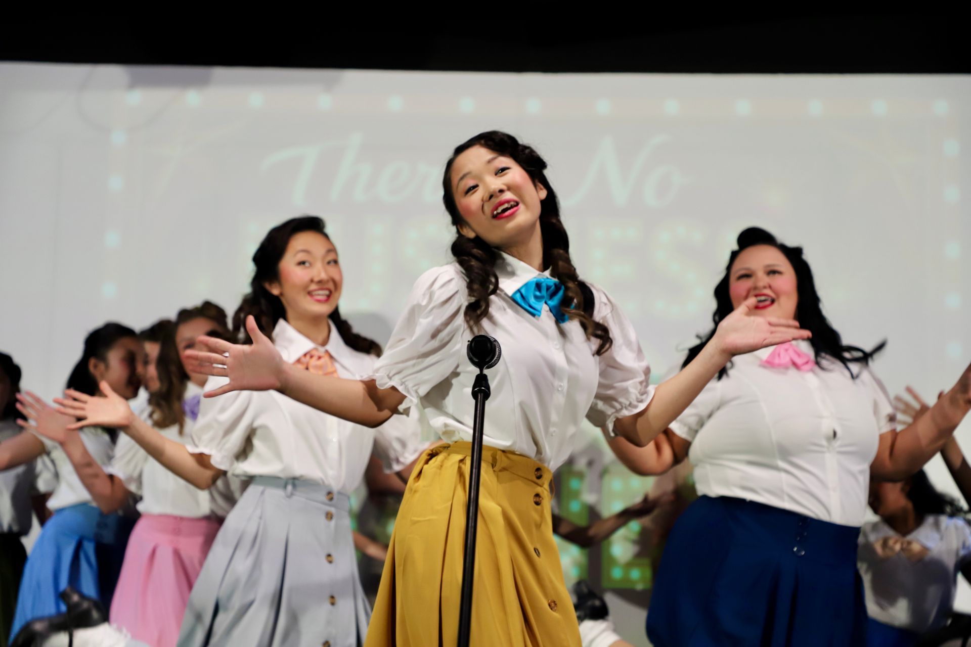 Dancers in white blouses and colorful skirts perform on stage with arms outstretched, for a theater performance.