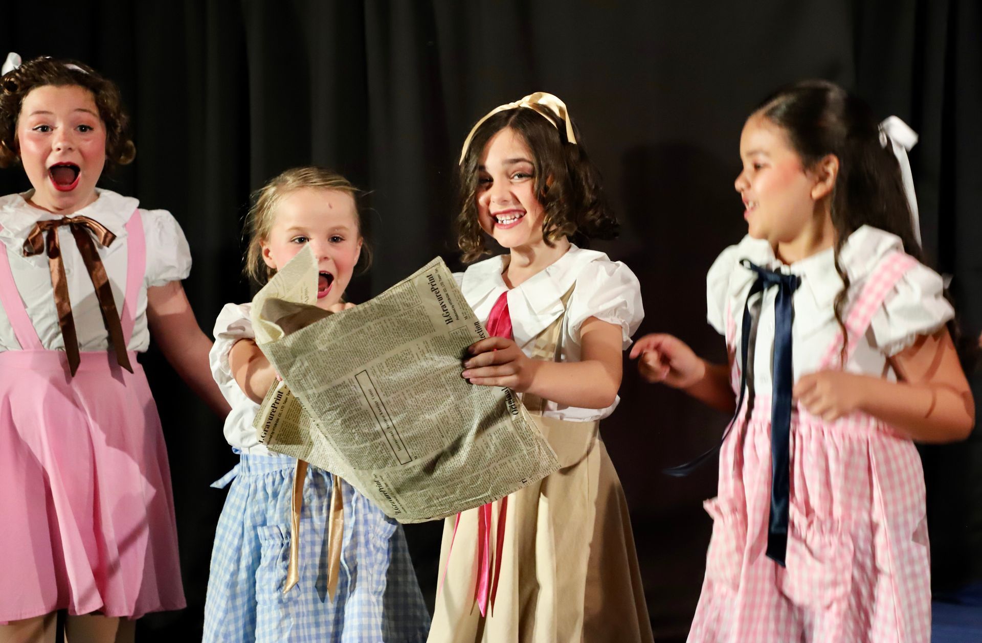 Children in pink and blue costumes singing and holding sheet music on stage for a theater performance.