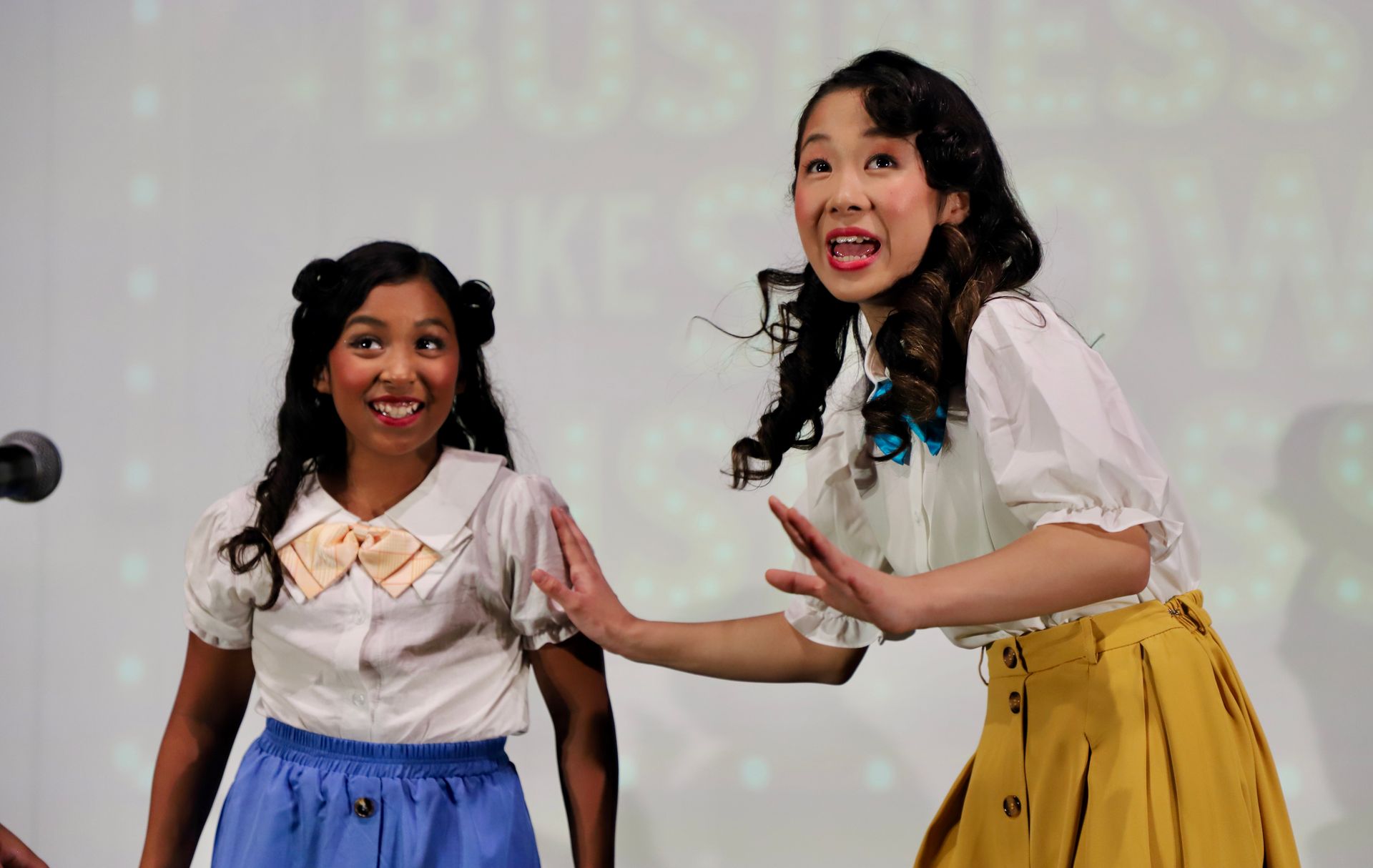 Two performers on stage, smiling and gesturing, in white tops with blue and yellow skirts for a theater performance.