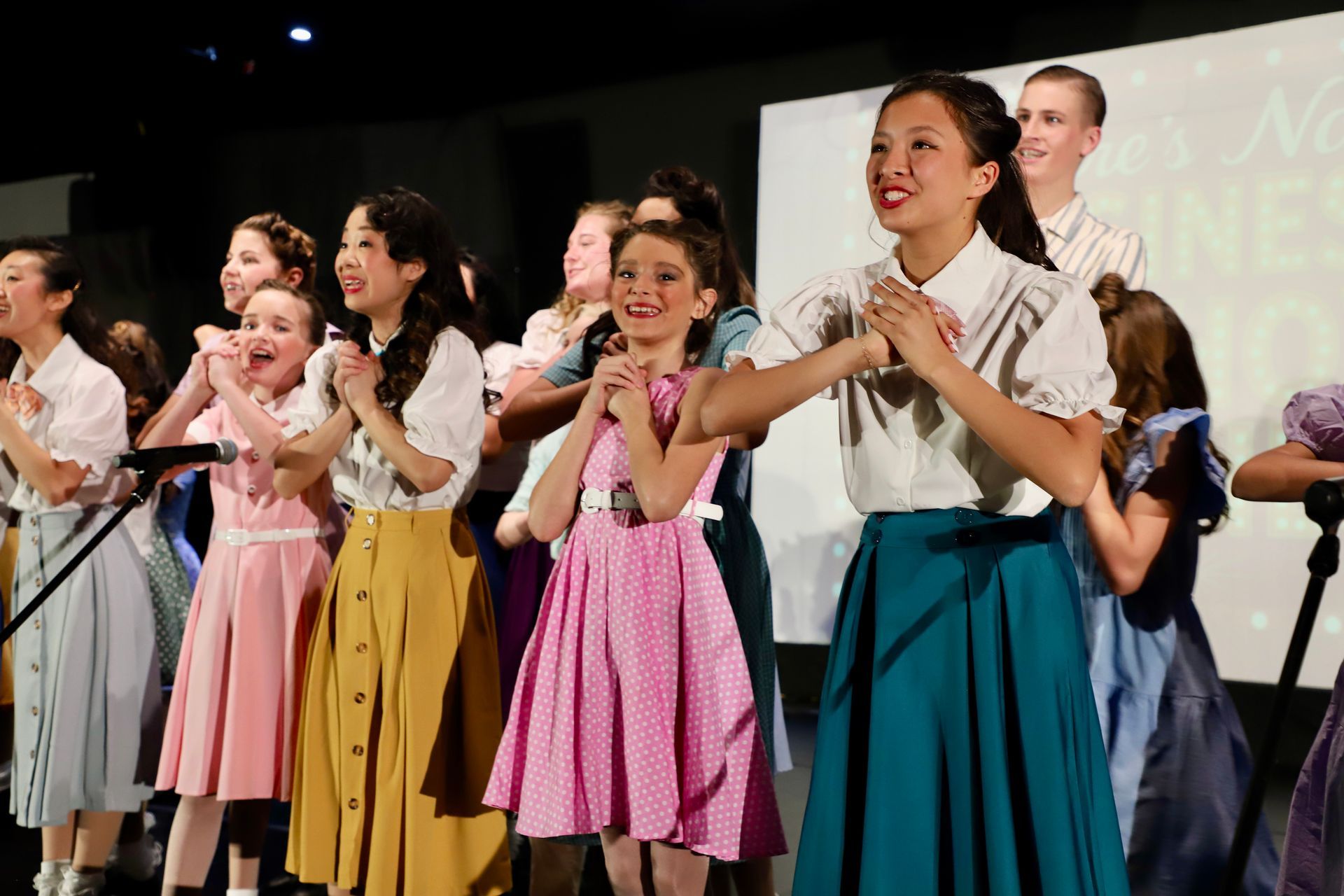 Children in colorful dresses sing and clap on stage during a performance for a theater performance.