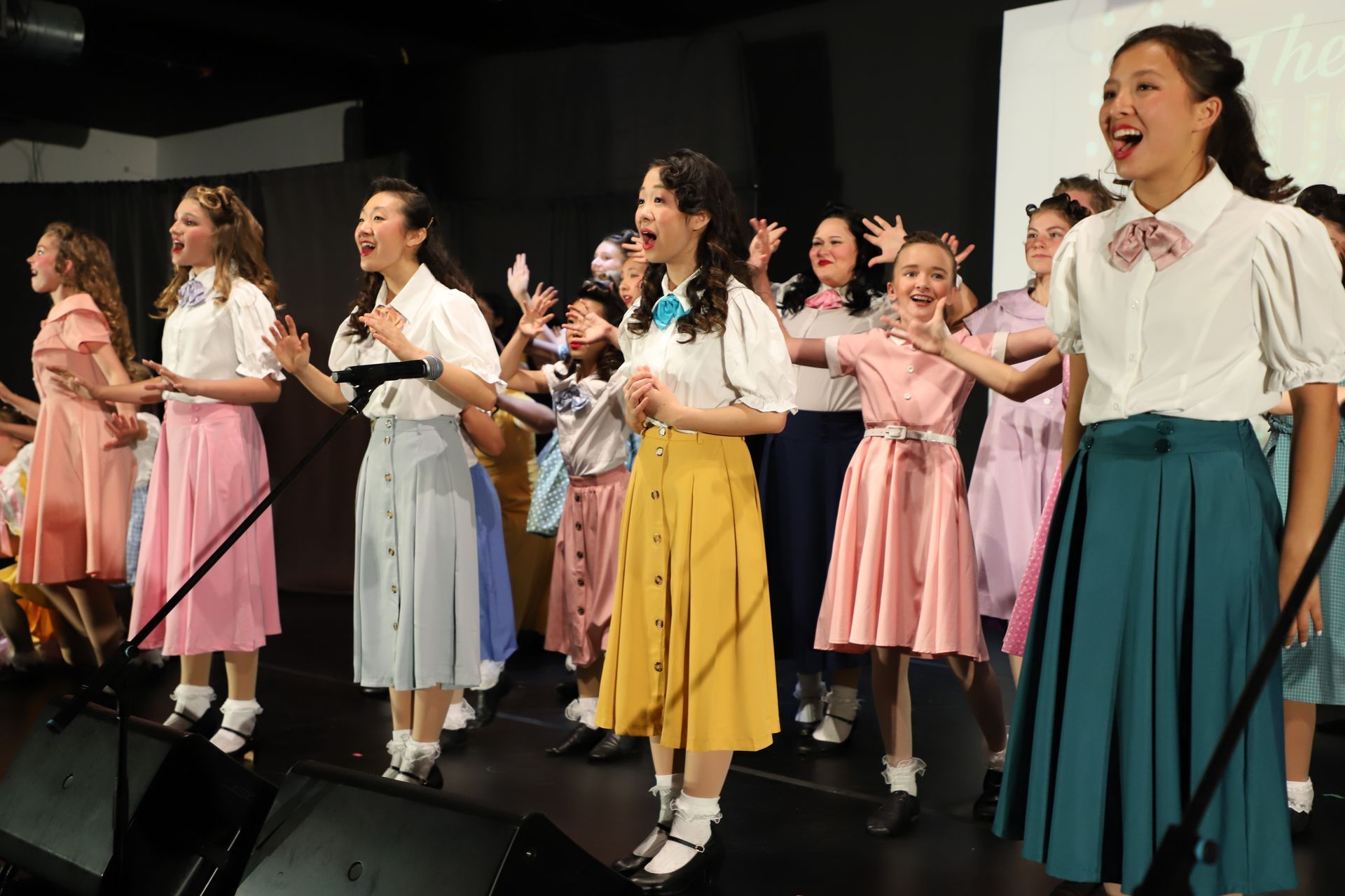 Choir group singing on stage, wearing colorful outfits and smiling with raised hands for a theater performance.