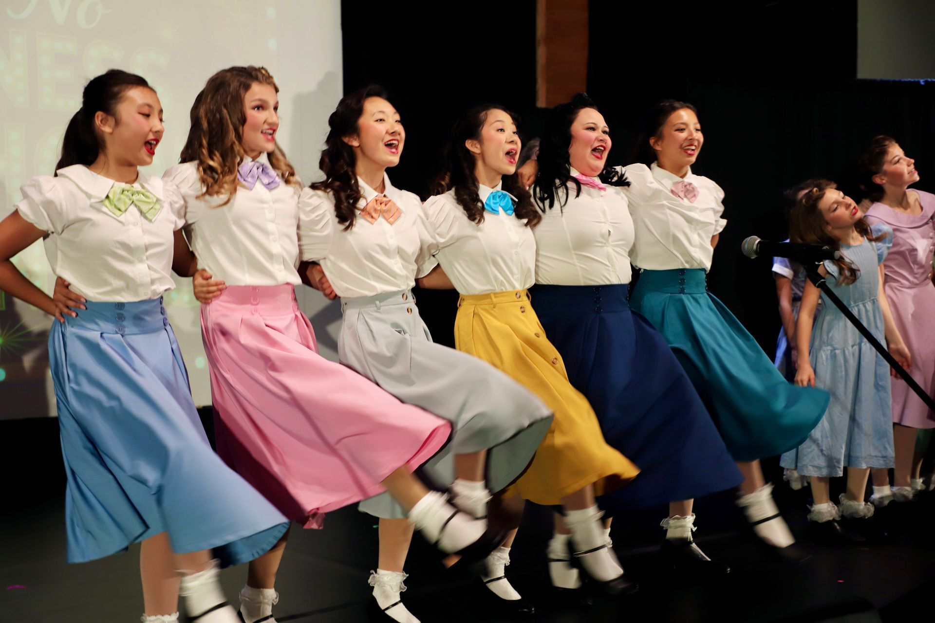 Six performers in colorful skirts sing and kick in unison onstage for a theater performance.