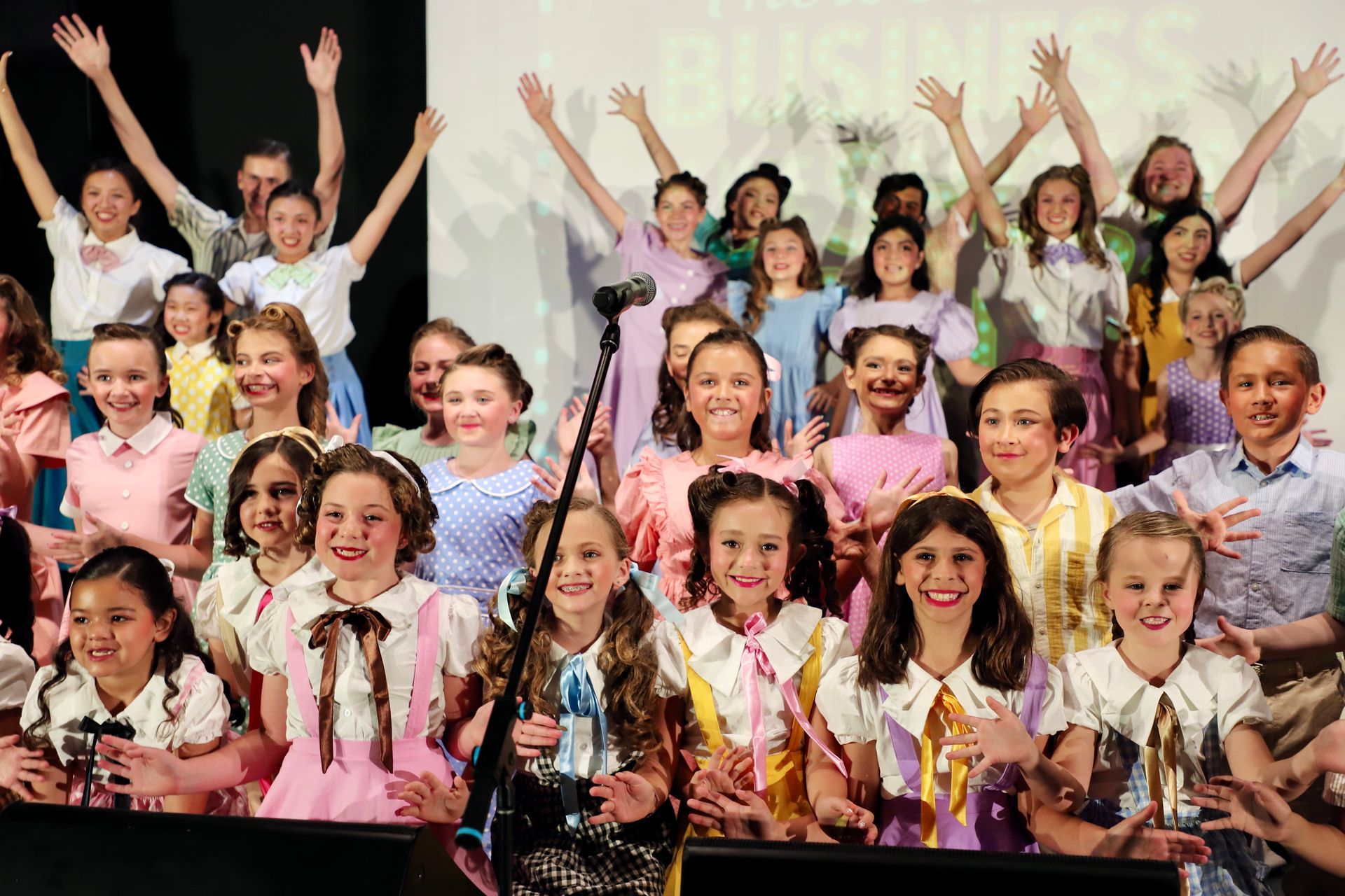 Large group on stage smiling and raising hands, wearing colorful costumes and holding trophies for a theater performance.