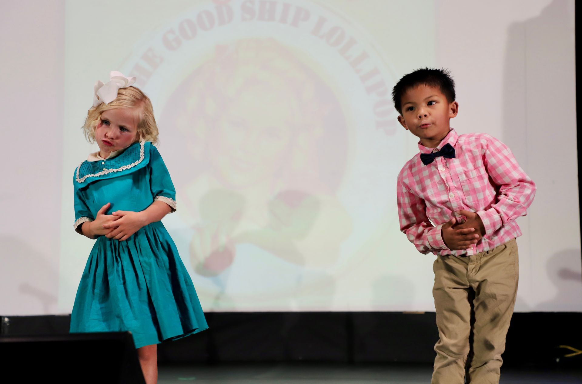 Two children stand on a stage, one in a teal dress and one in a pink shirt for a theater performance.