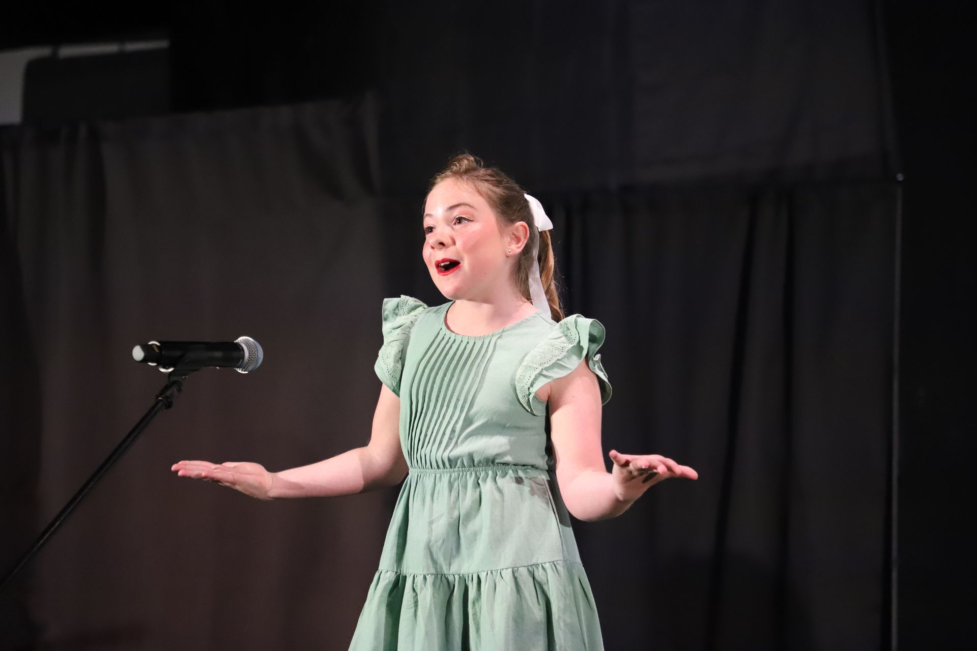 Child singing onstage in a green dress beside a microphone, arms outstretched for a theater performance.