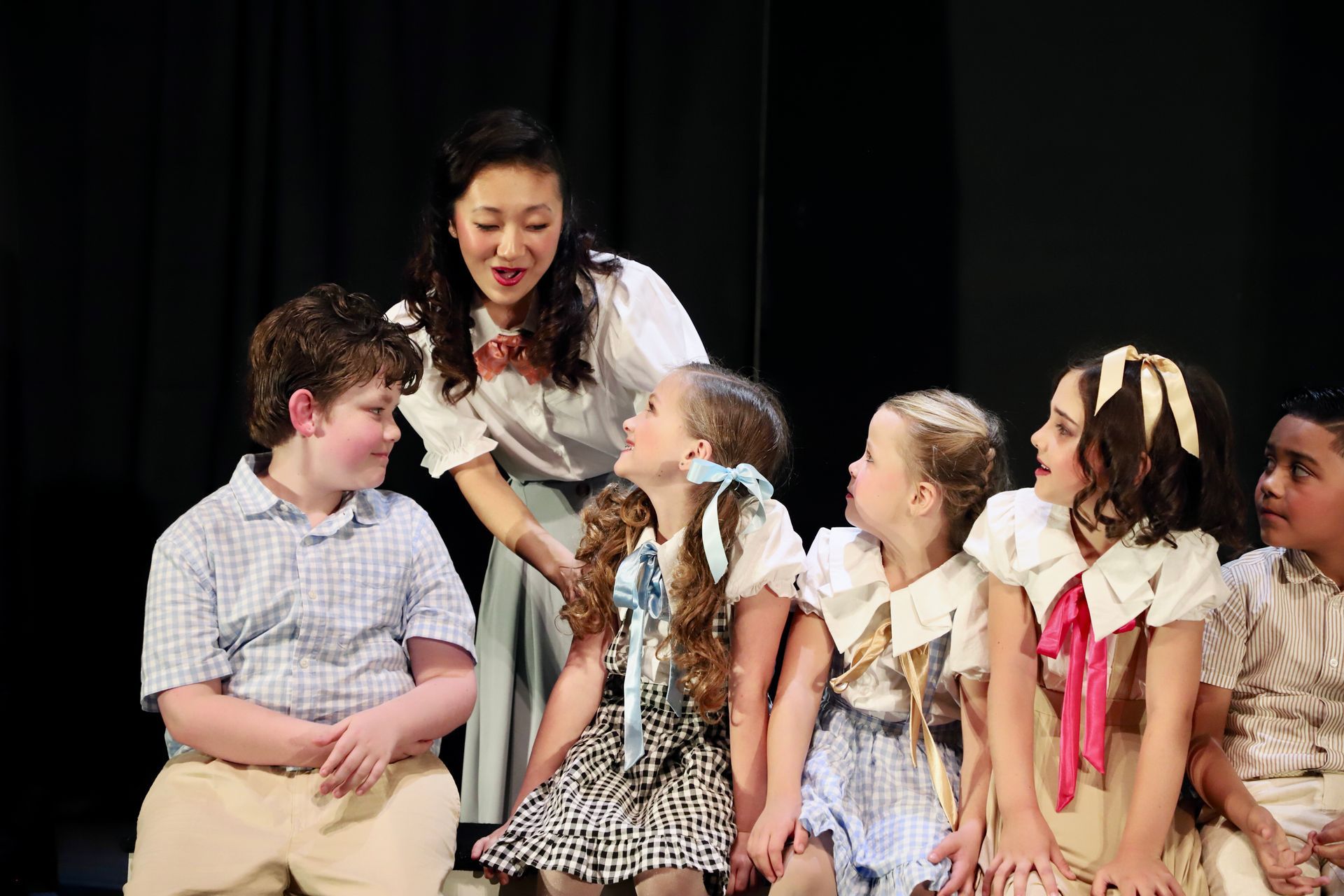 Teacher kneels beside seated children onstage, speaking to them under warm stage lights for a theater performance.