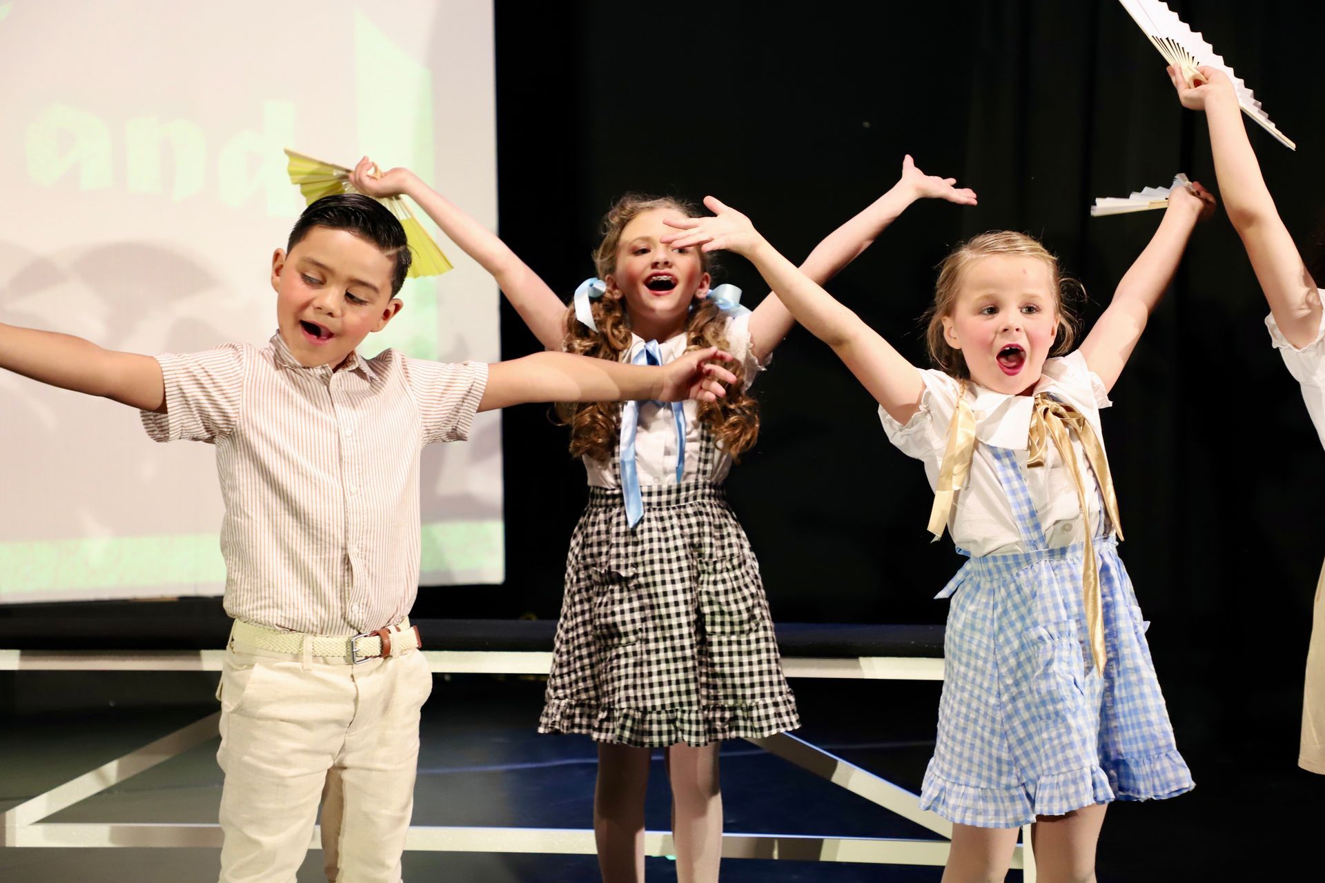 Three children on stage with arms raised, smiling and singing in colorful outfits for a theater performance.