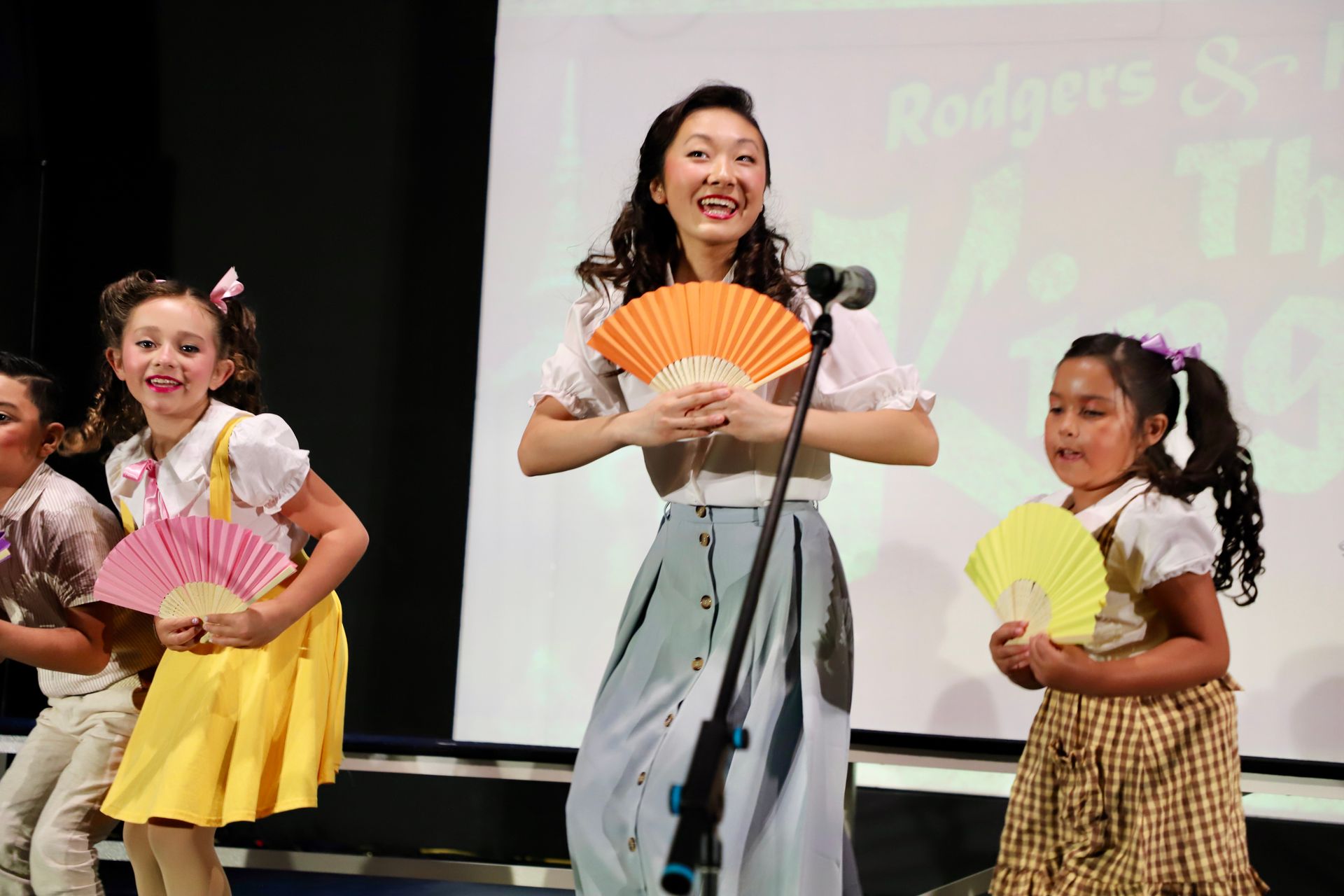 Three performers on stage hold colorful fans during a dance performance for a theater performance.
