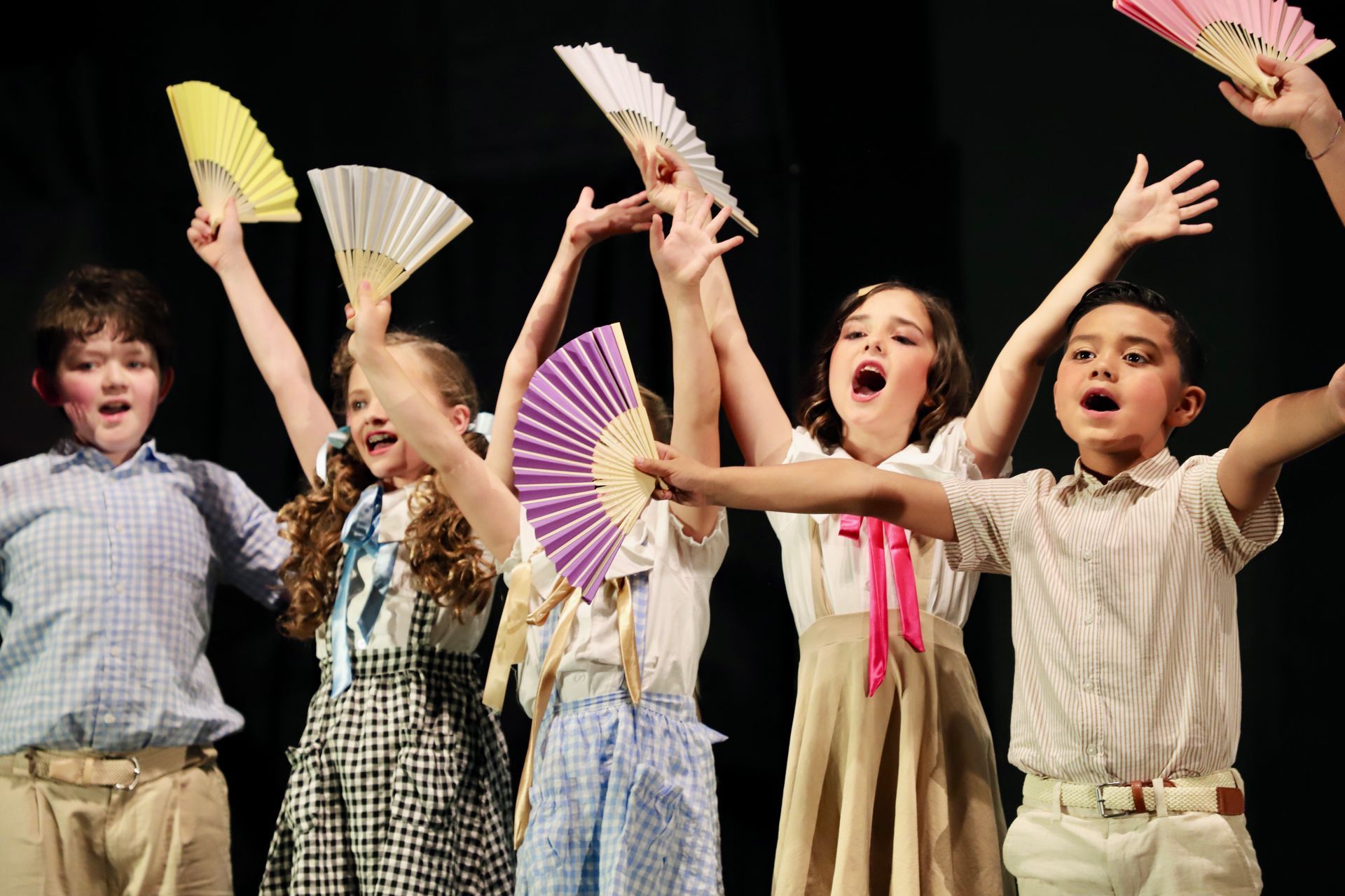 Children on stage waving paper fans, smiling and cheering under warm lights for a theater performance.