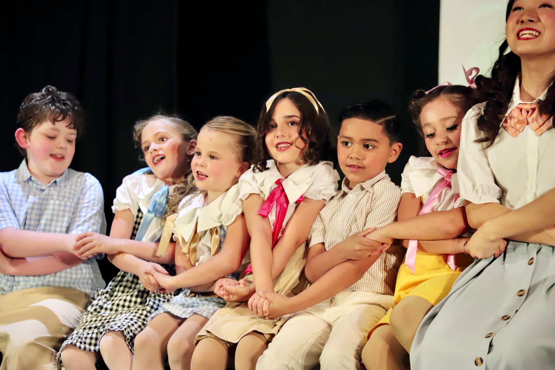 Children seated on stage, smiling and laughing in colorful outfits for a theater performance.